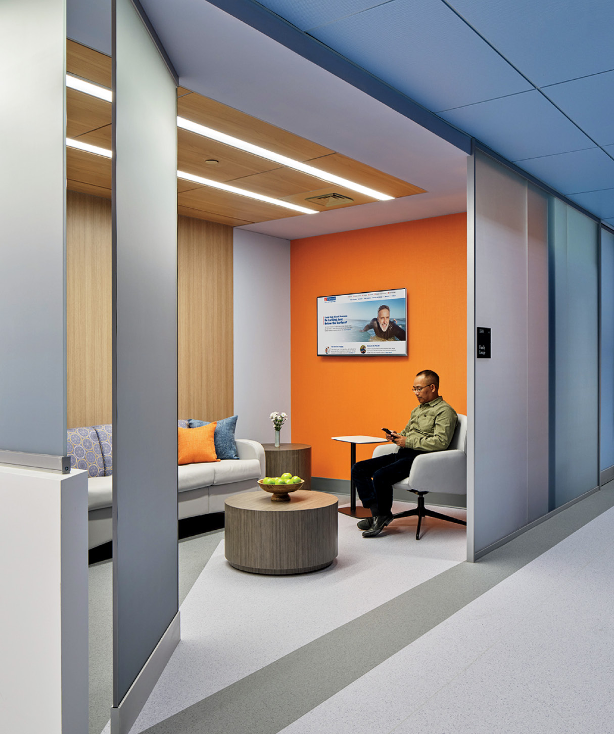 A man sits in a modern waiting area with orange accent wall, gray sofa, round table with fruit bowl, and a wall-mounted TV displaying a news segment.