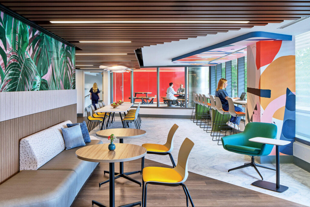 Modern office lounge with colorful decor, tables, chairs, and people working or talking. Large windows and vibrant wall art create a bright, open atmosphere.