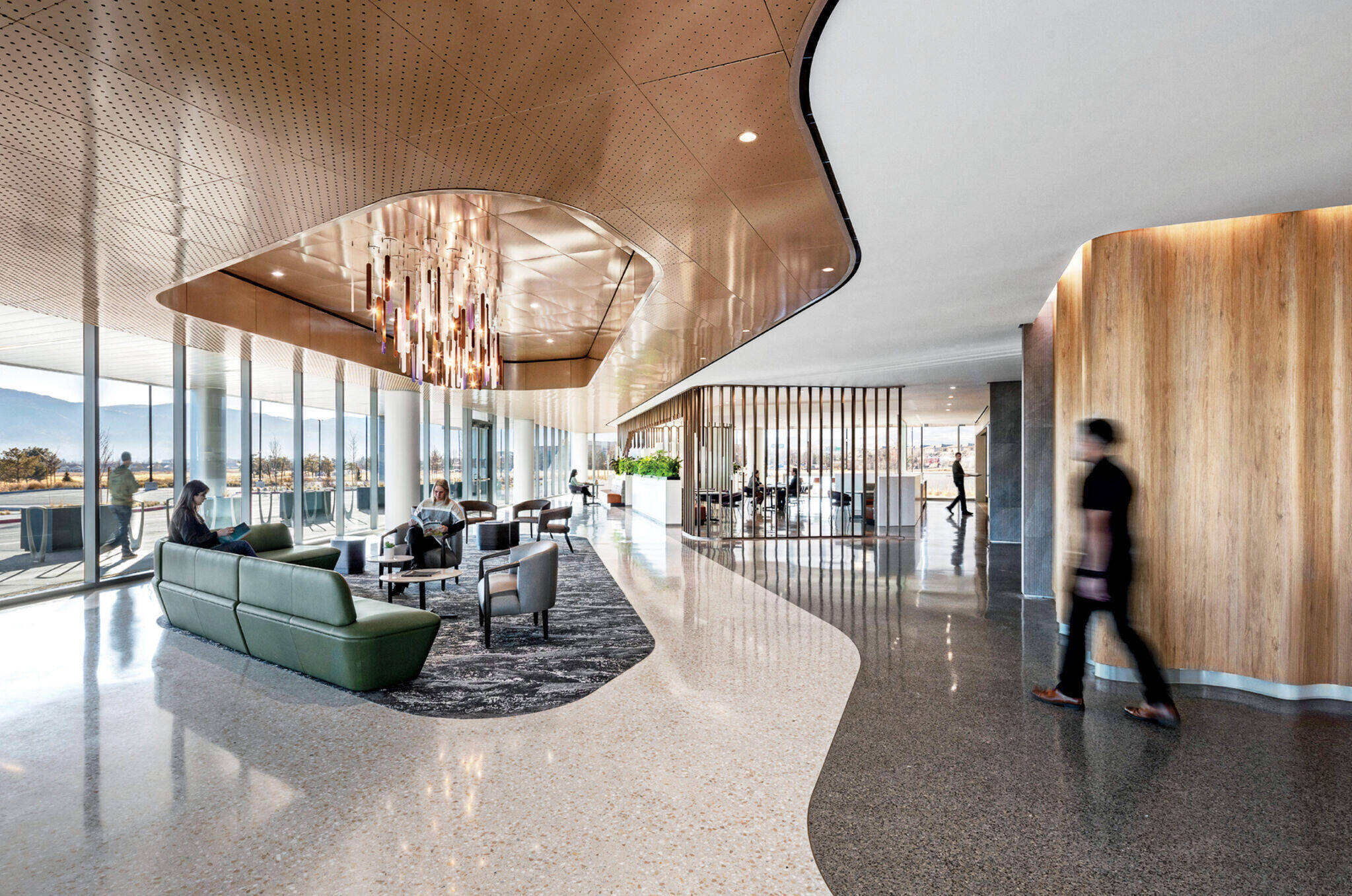 Modern office lobby with curved wood ceiling, contemporary lighting, green seating area, large windows, and several people walking or sitting.