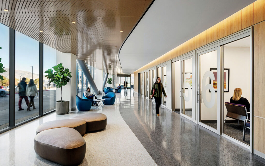 Modern office lobby with glass-walled rooms, people sitting and walking, large windows, cushioned seats, indoor plants, and natural light.