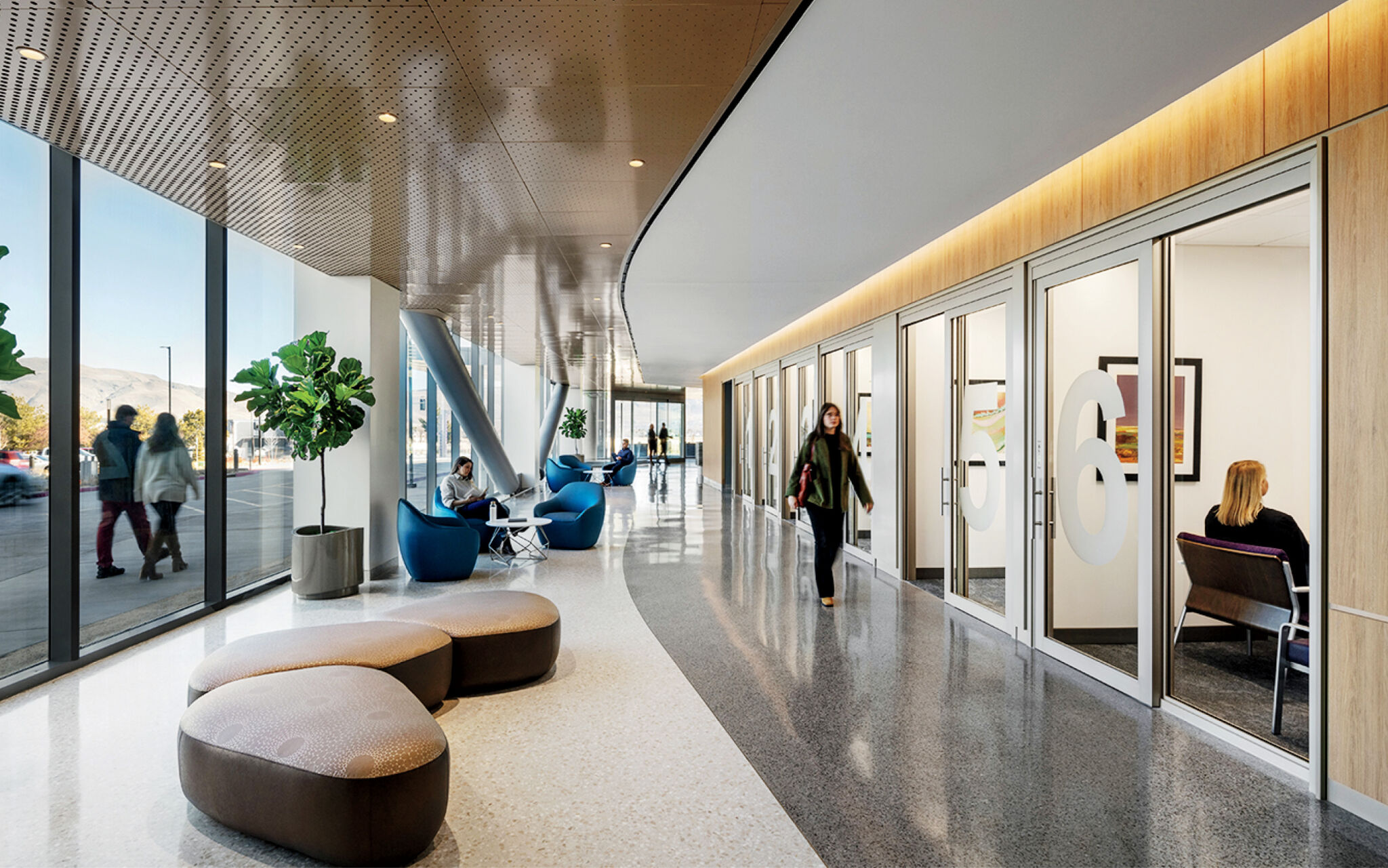 Modern office lobby with glass-walled rooms, people sitting and walking, large windows, cushioned seats, indoor plants, and natural light.