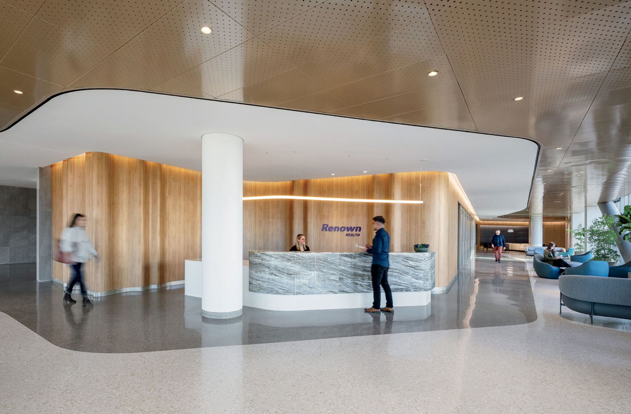 Modern reception area with a marble front desk, a person speaking to the receptionist, and others walking or sitting in a spacious, well-lit lobby.