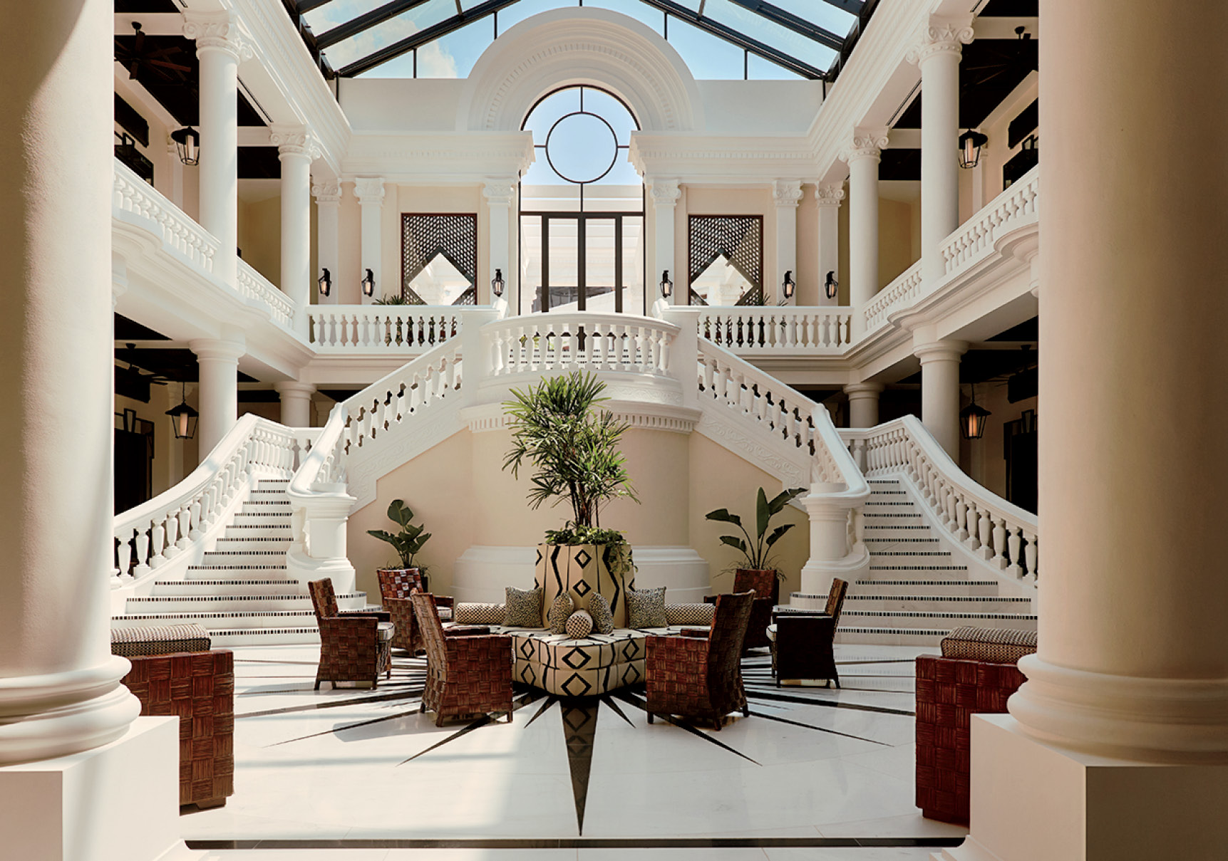 Grand hotel lobby with high glass ceiling, twin sweeping staircases, white columns, central fountain, potted plants, and wicker chairs arranged around a round table on a marble floor.