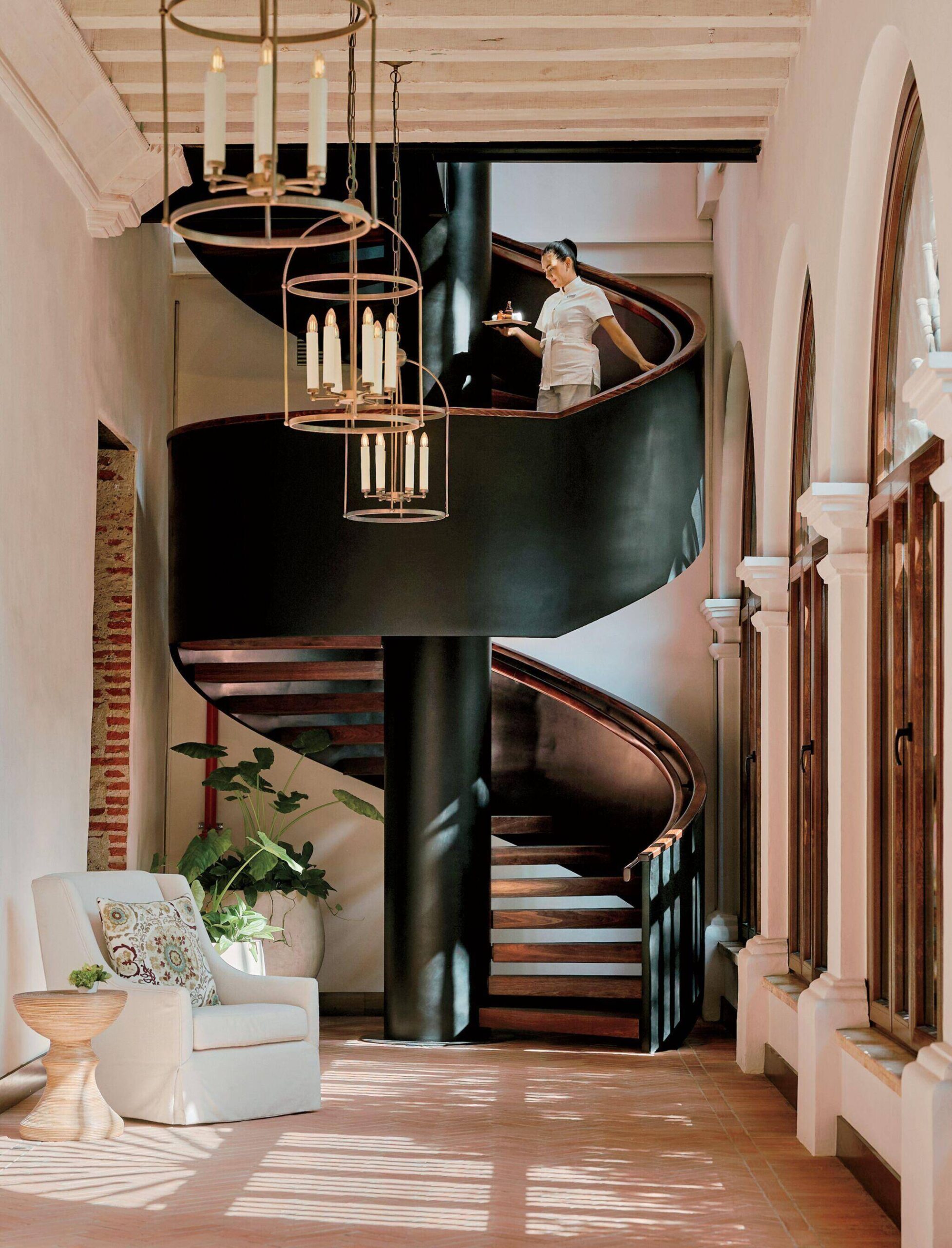 A person descends a black spiral staircase in a sunlit room with arched windows, a white armchair, potted plants, and a hanging chandelier.