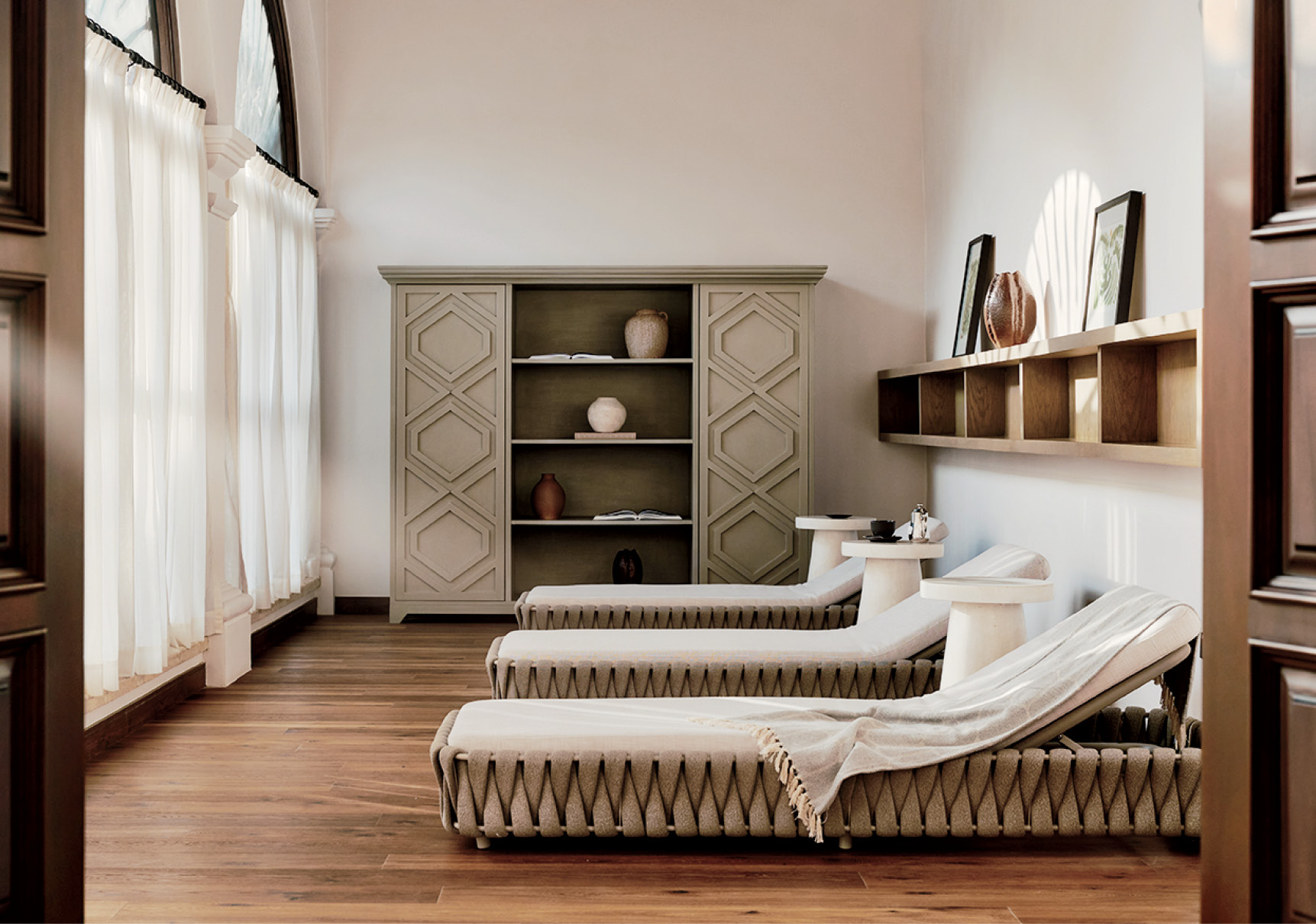 Three cushioned lounge chairs with rolled towels face a cabinet and wall shelves in a sunlit room with wooden floors and large curtained windows.