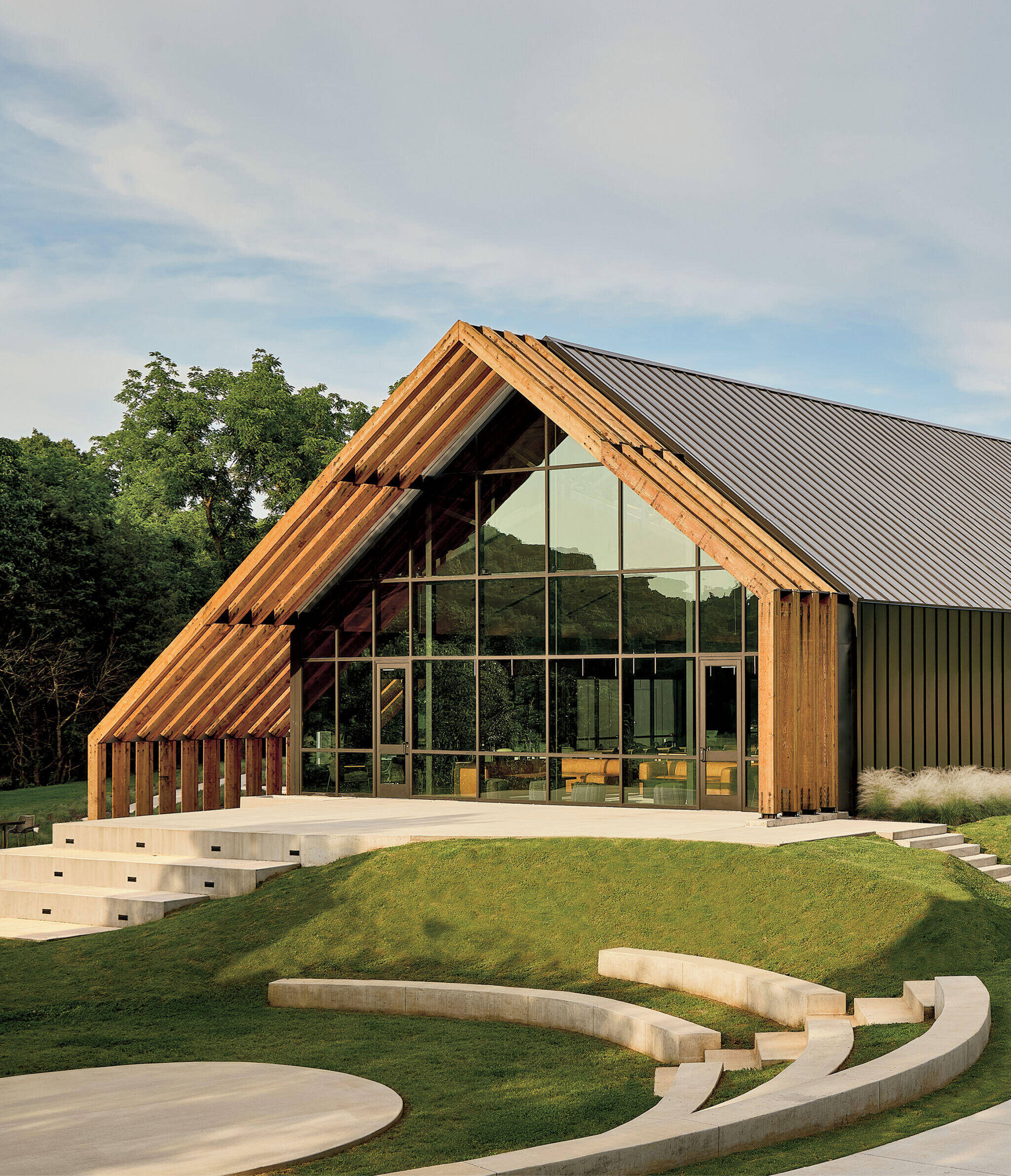 Modern building with large glass windows and wooden beams, surrounded by green landscaping and tiered concrete seating steps.