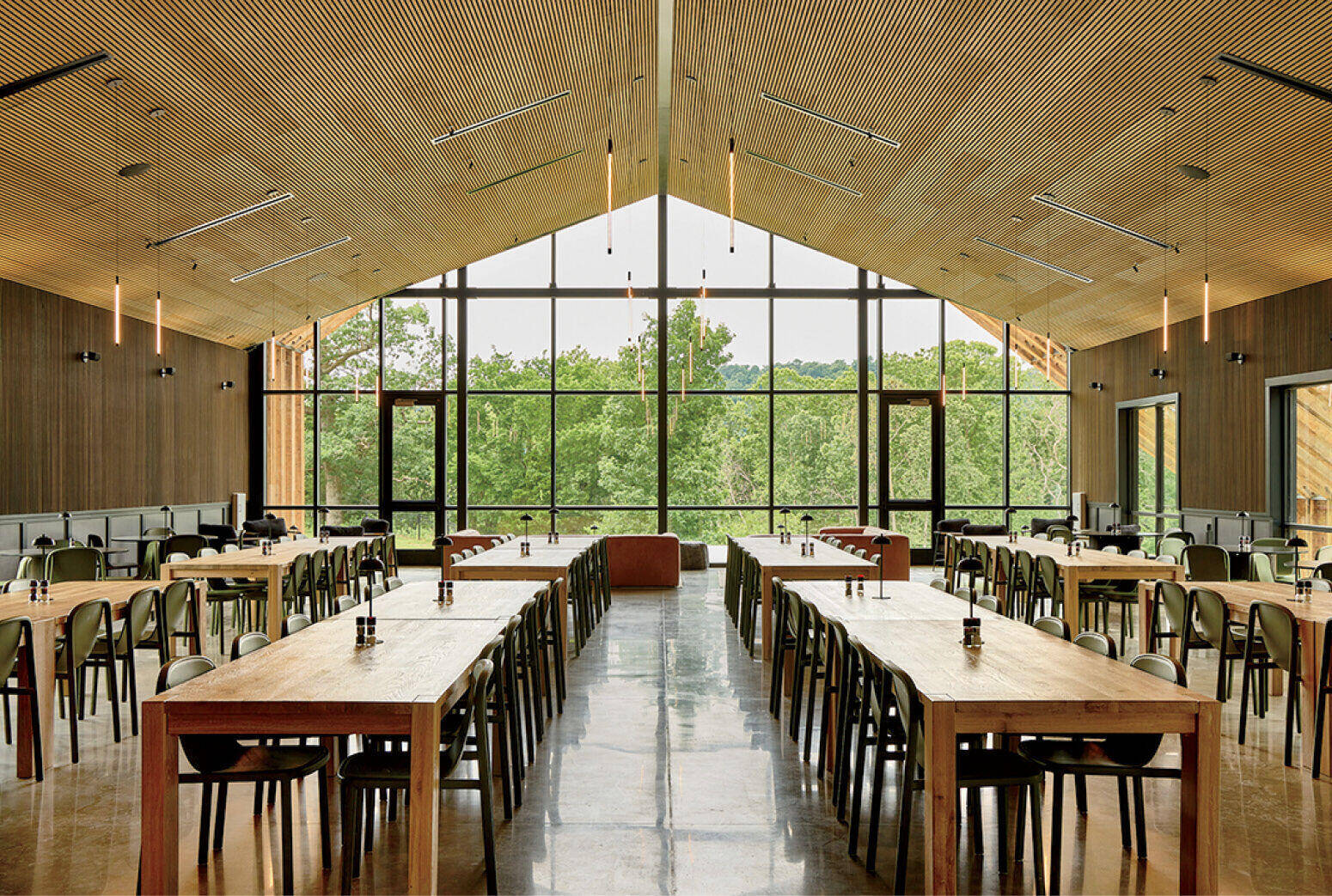 Spacious dining hall with wooden tables, metal chairs, tall windows, and a wood-paneled ceiling overlooking green trees outside.