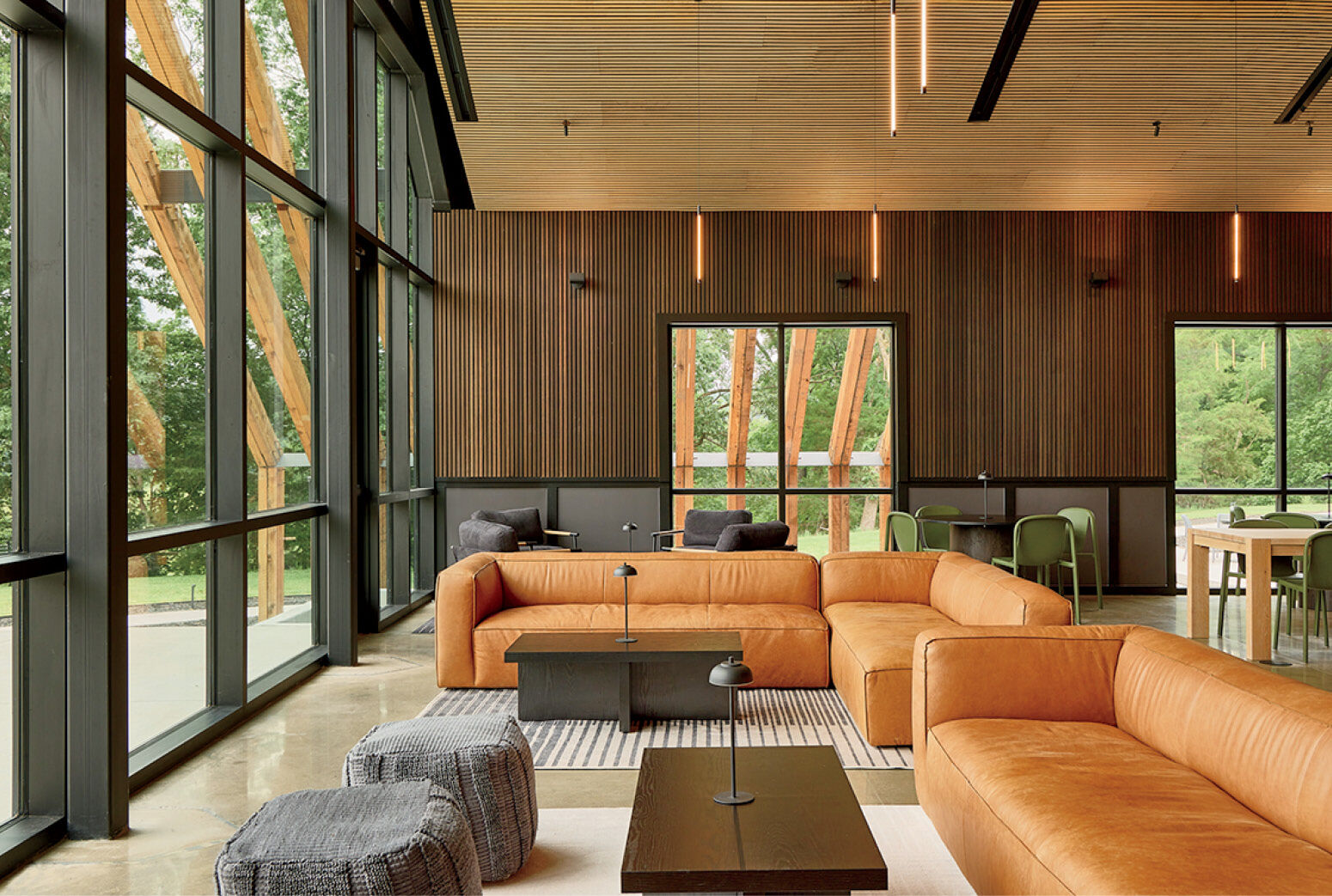 Modern lounge with tan leather sofas, dark wood walls, large windows, and minimalist decor; trees and greenery visible outside.