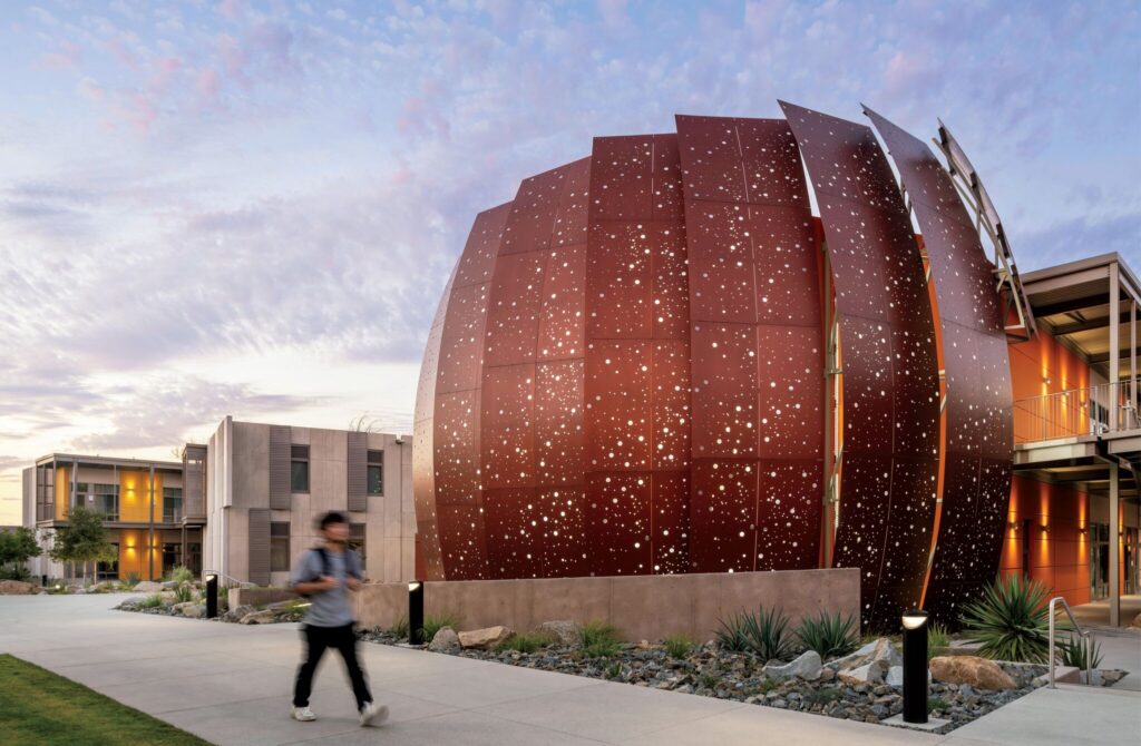A person walks past a large, spherical building with rust-colored, perforated panels on a modern campus at dusk.