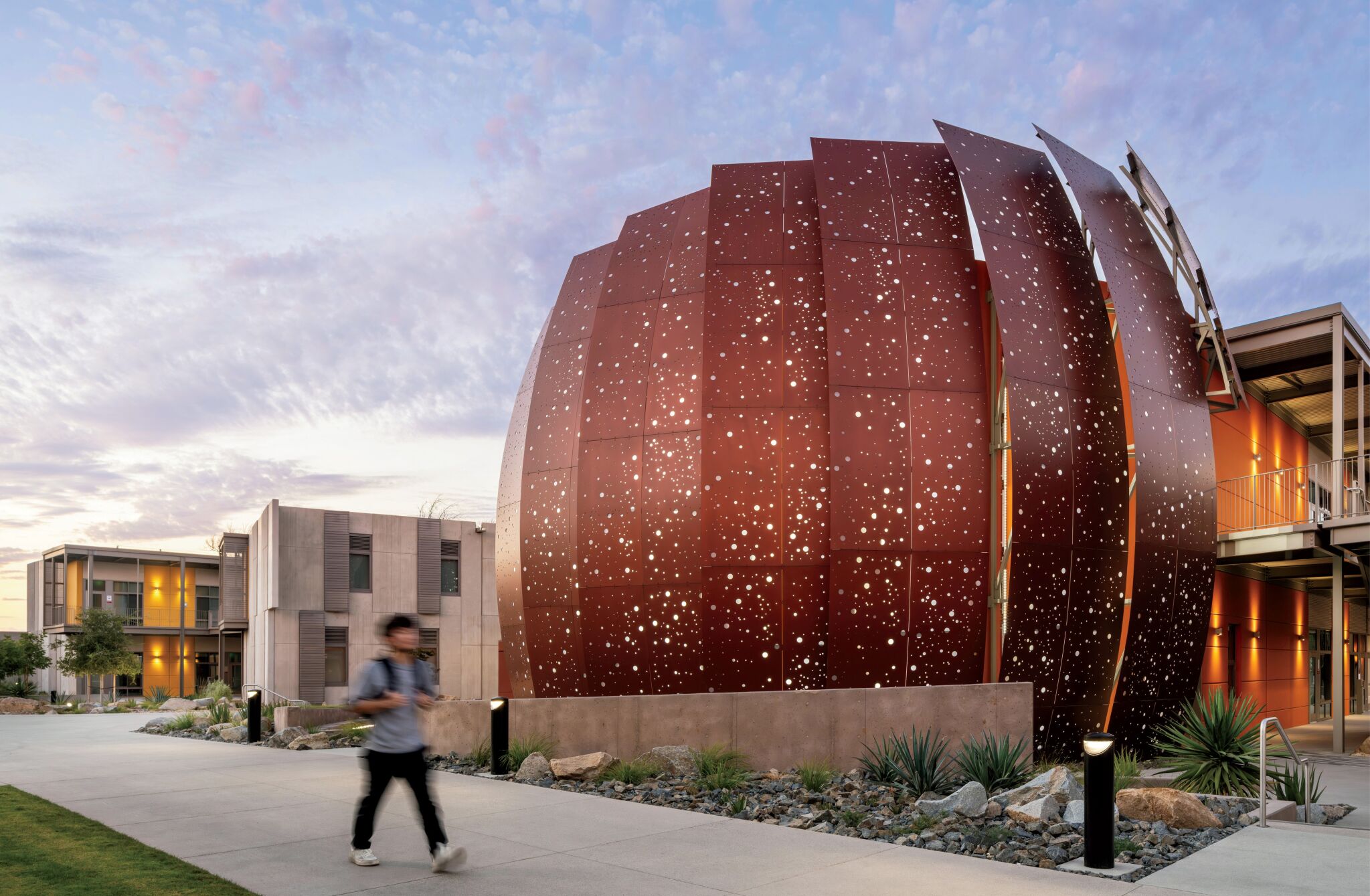 A person walks past a large, spherical building with rust-colored, perforated panels on a modern campus at dusk.