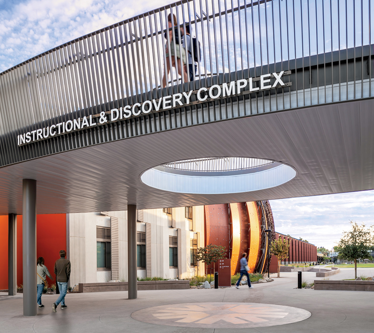 People walk near a modern building labeled “Instructional & Discovery Complex,” featuring a circular opening in the overhang and a pedestrian bridge above.