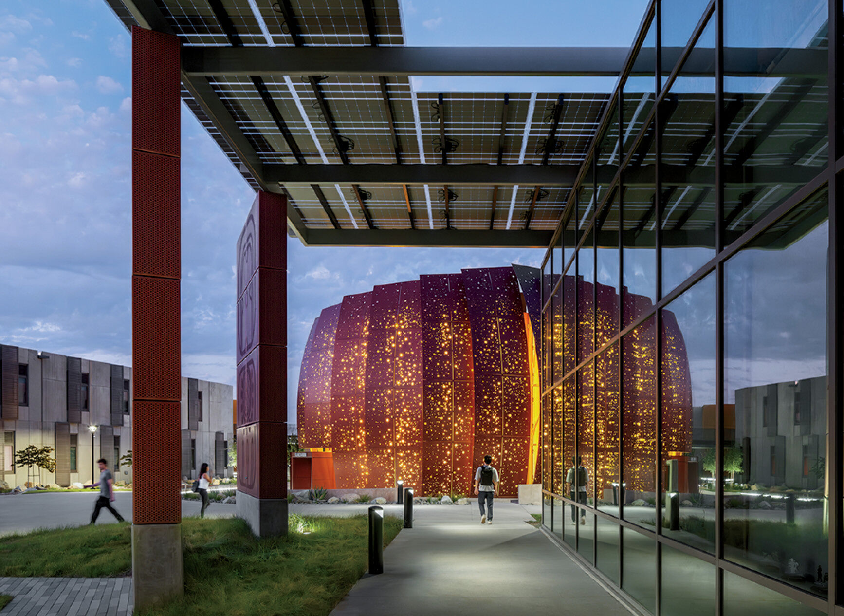 People walk near a modern building with an illuminated, dome-shaped structure featuring numerous small holes, viewed through a glass corridor at dusk.
