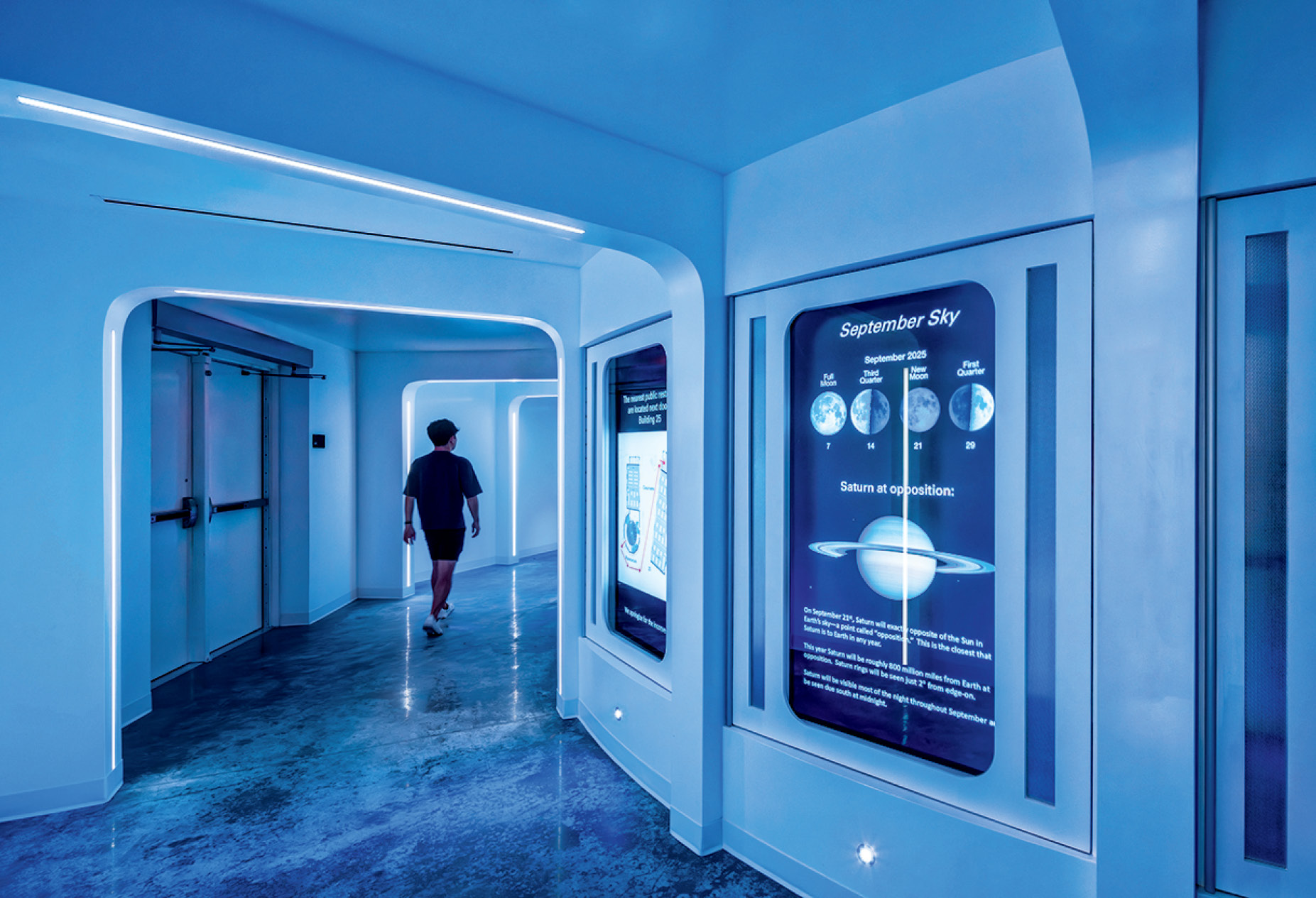 A person walks down a futuristic hallway with blue lighting, glossy floors, and large digital screens displaying astronomical information.
