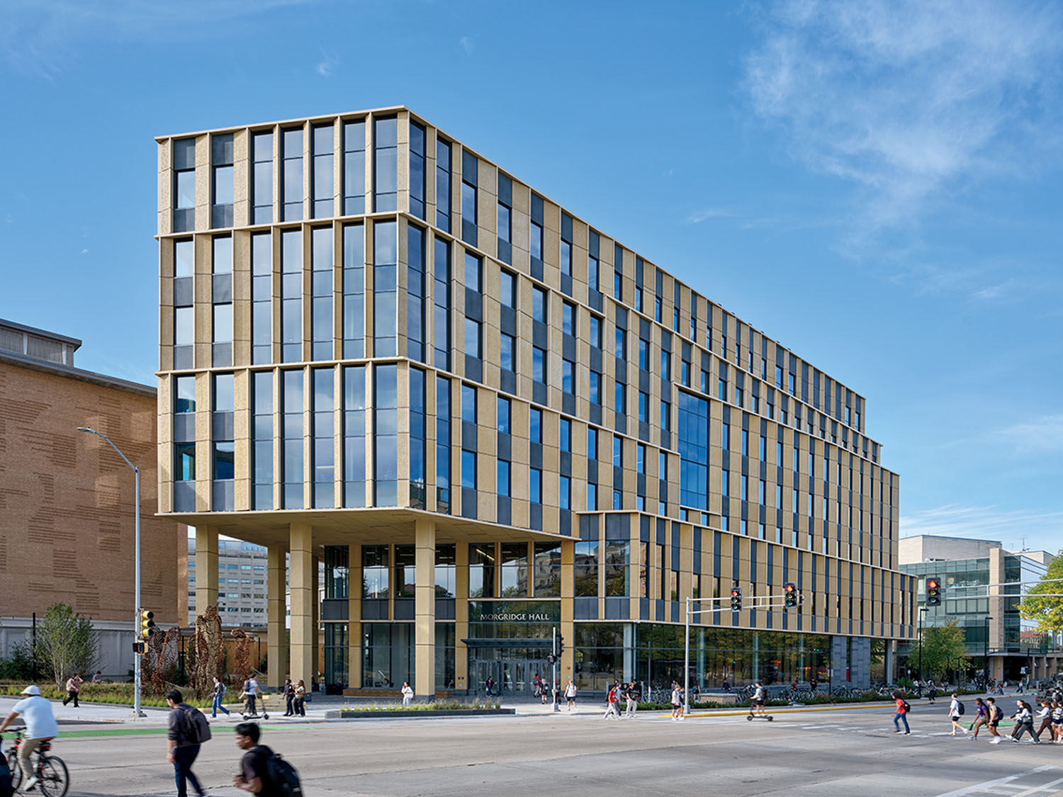 A modern, multi-story glass and stone building with large windows, situated on a busy street corner with pedestrians and cyclists crossing.