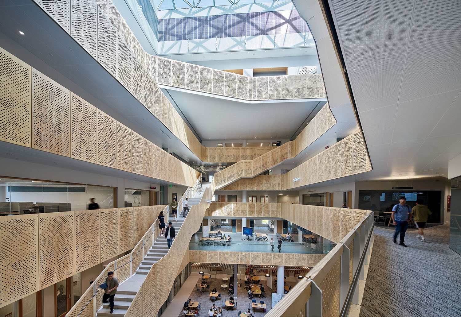 Modern atrium with multiple floors, open staircases, patterned railings, skylight ceiling, and people walking and sitting in communal areas below.