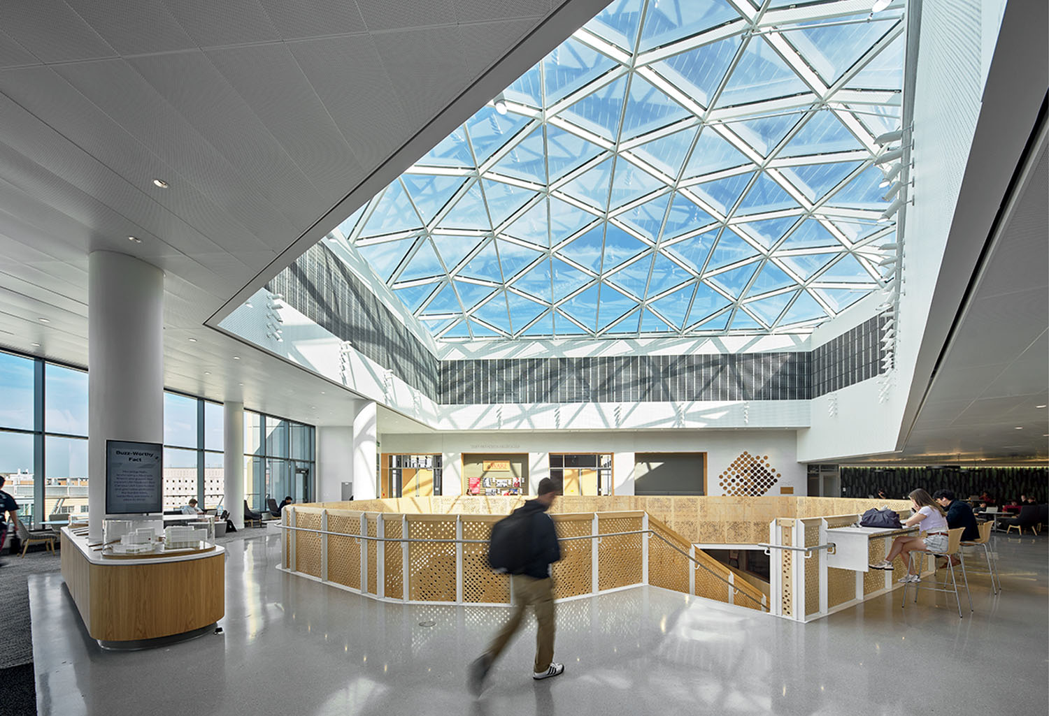 Spacious modern atrium with geometric glass ceiling, natural light, people sitting at tables, and one person walking in the center.