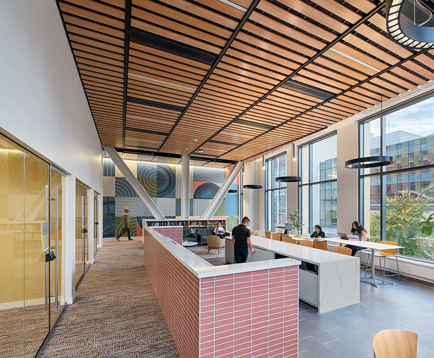 Modern office lounge with large windows, wooden ceiling panels, people working at tables, and a central counter area. Natural light fills the space.
