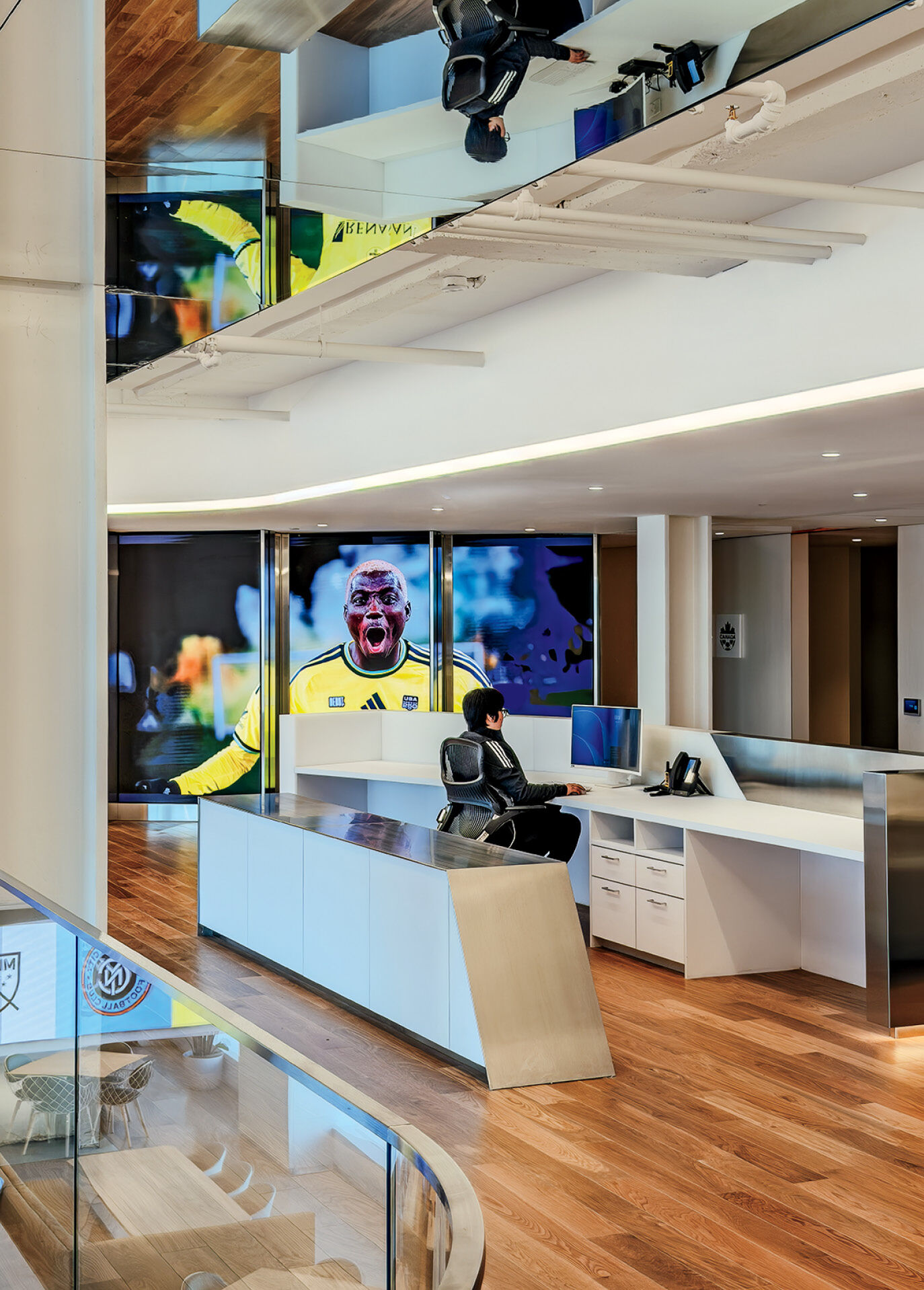 Modern office lobby with a receptionist at a white desk, wooden floors, a large sports mural, and a mirrored ceiling reflecting the space.