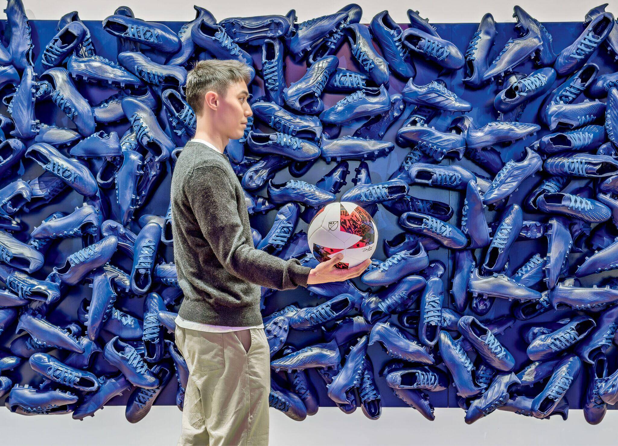 A man in a gray sweater holds a soccer ball, standing in front of a wall covered with blue soccer cleats arranged in a dense, patterned display.