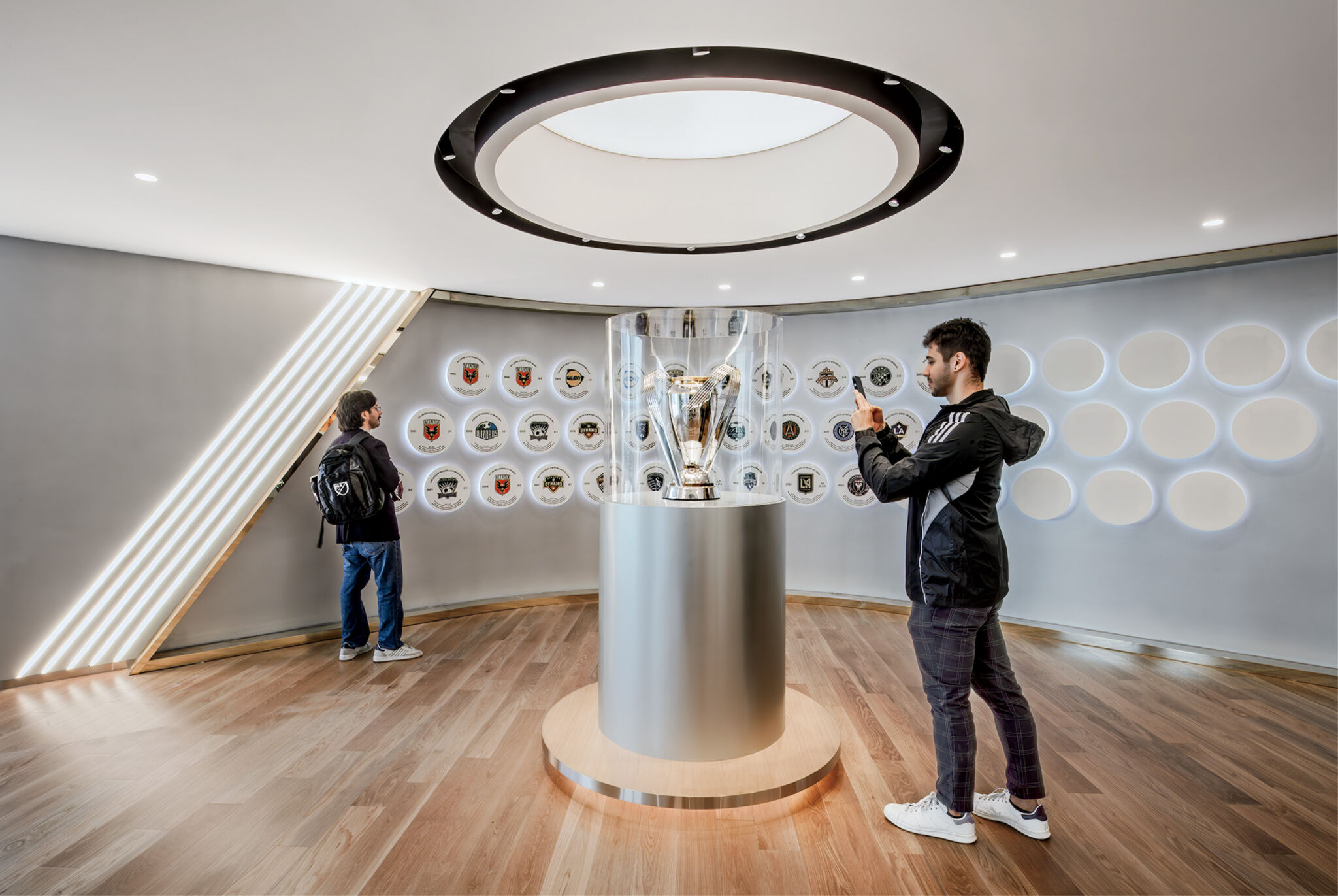 Two people observe and photograph a large trophy displayed in a glass case in a modern, well-lit room with wooden floors and football club logos on the wall.