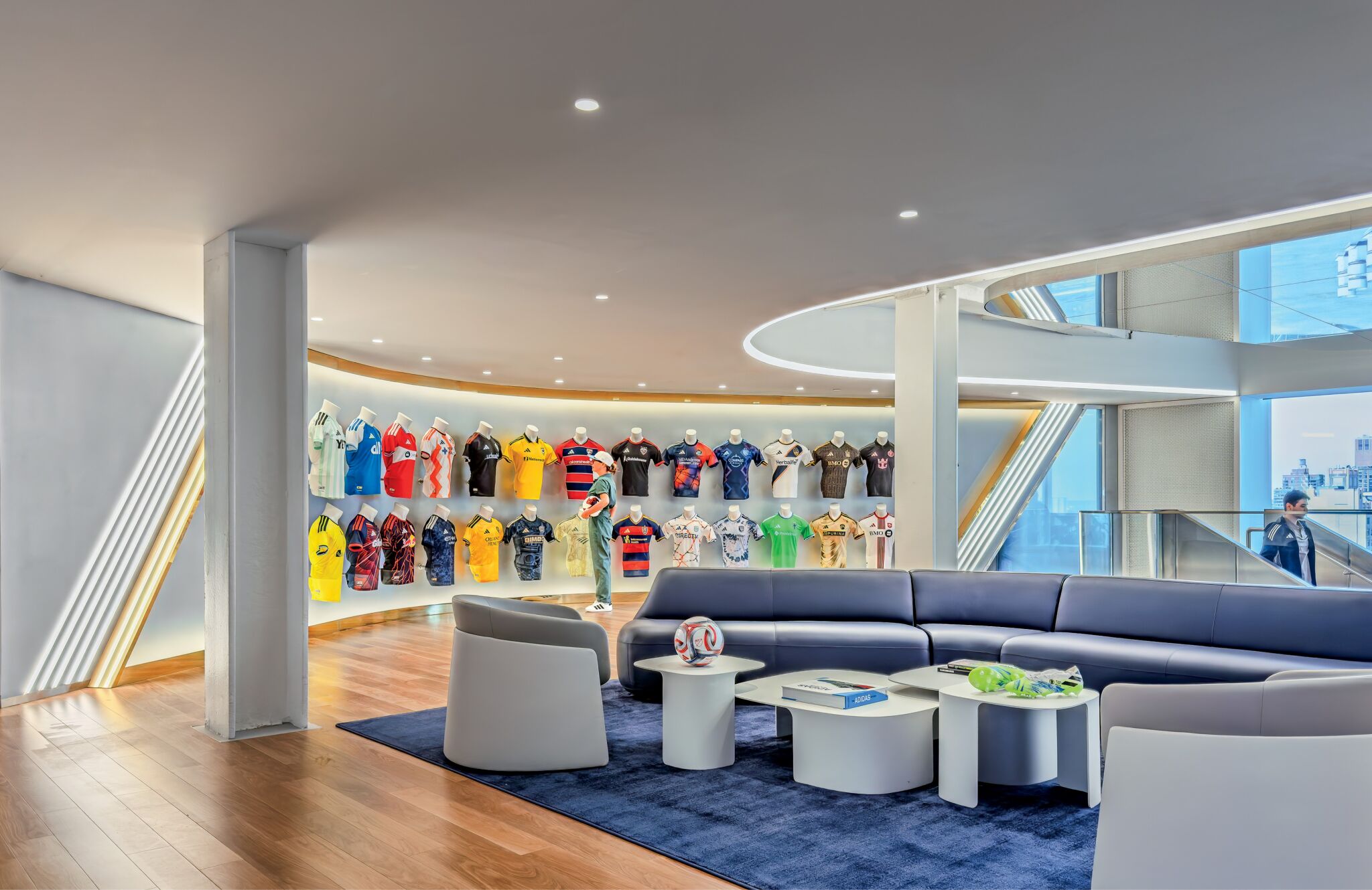 Modern lounge with curved blue couches and a display wall featuring colorful sports jerseys. Coffee tables in the center hold balls and memorabilia. Large windows provide city views.
