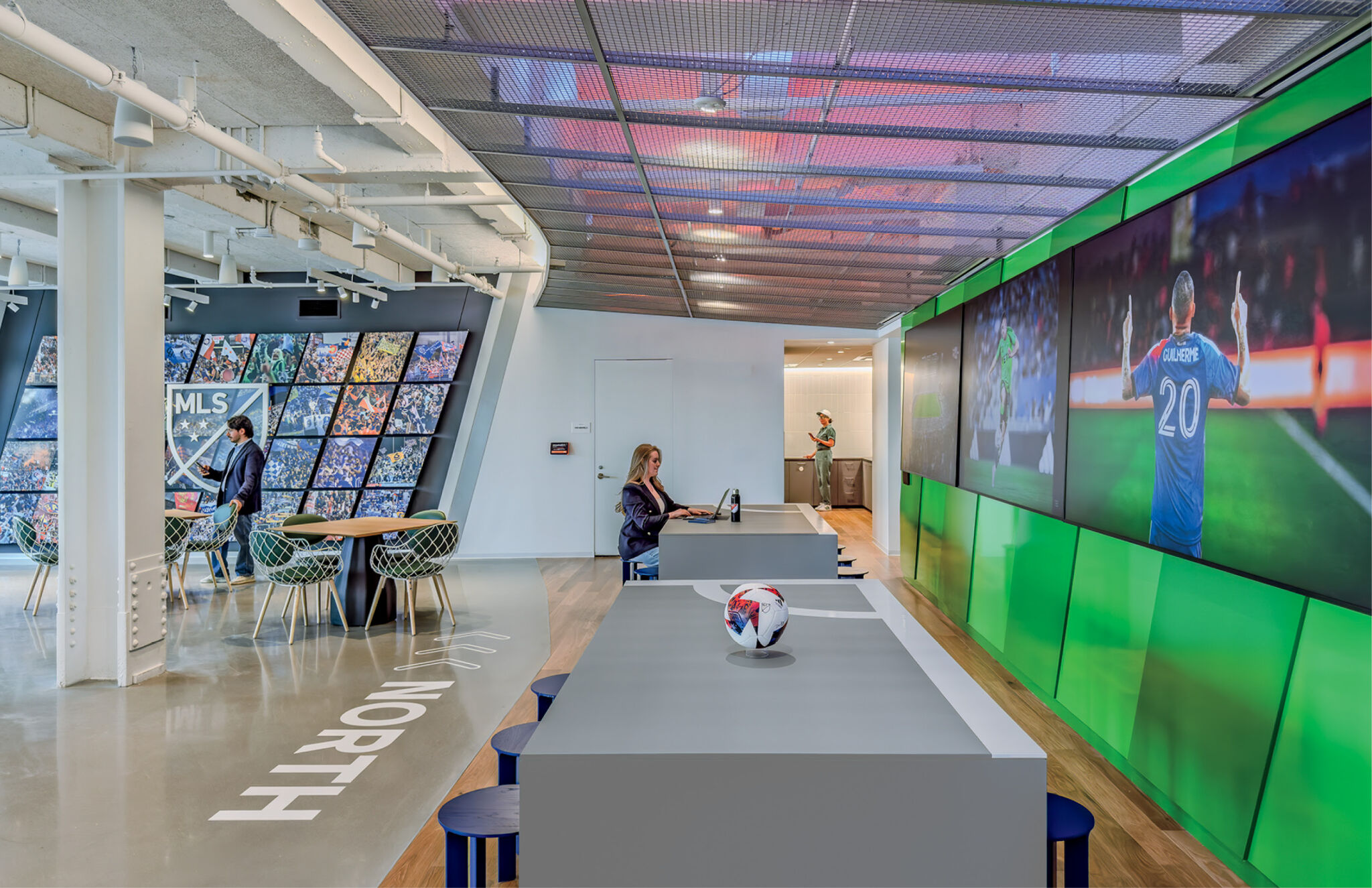 Modern office space with large video screens showing soccer, a woman working at a table, another person near a wall display, and a soccer ball on a central table.