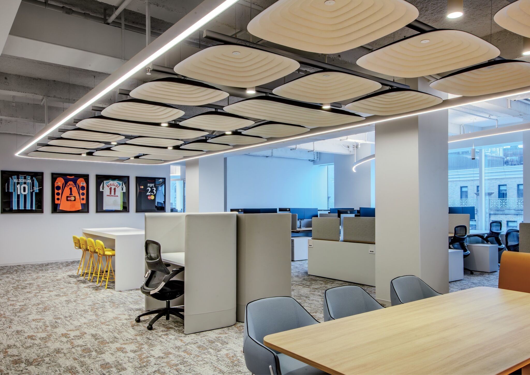 Modern open office space with desks, chairs, a wooden table, yellow stools at a counter, and sports jerseys displayed on the wall. Unique ceiling panels and large windows provide natural light.