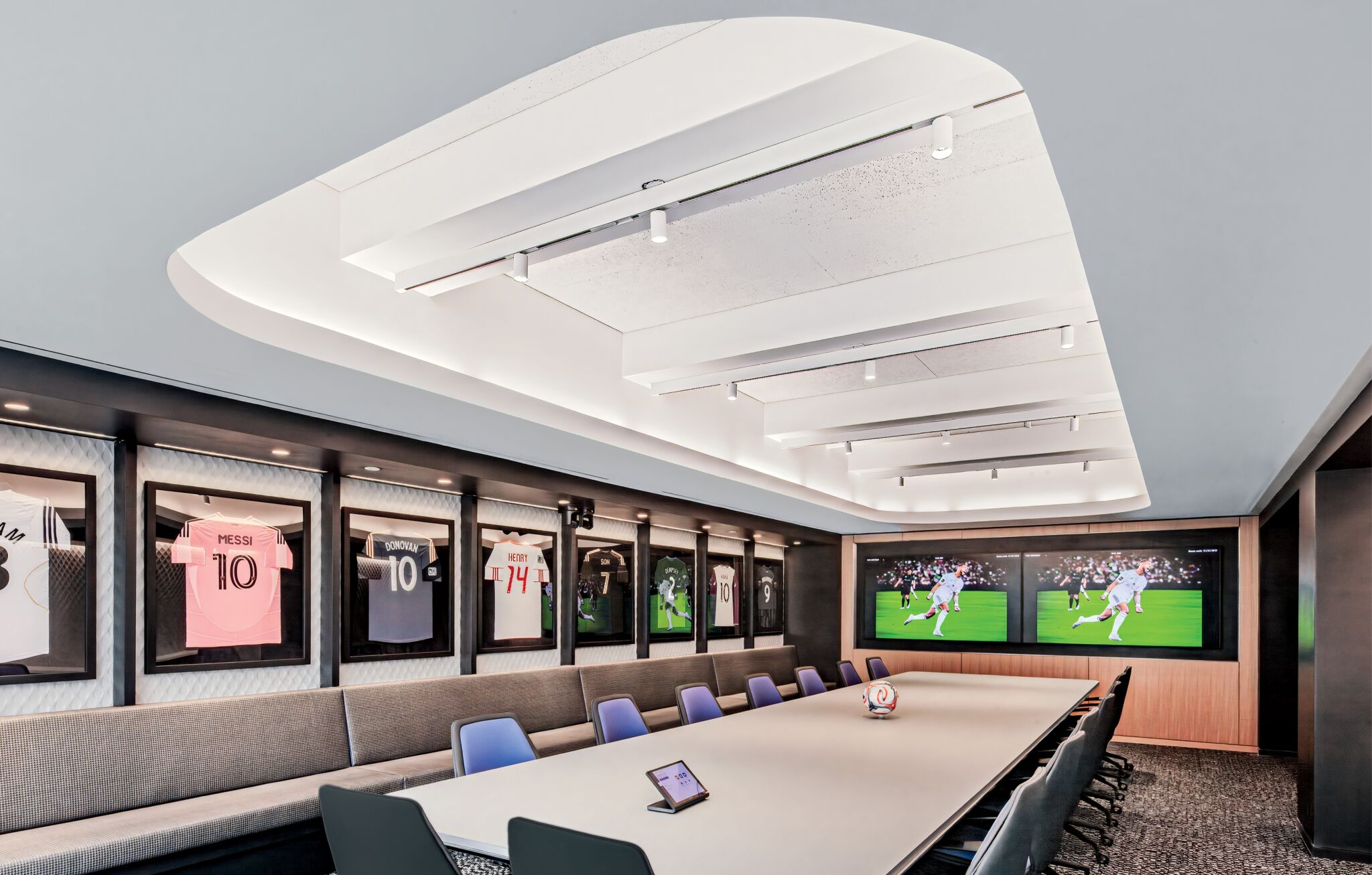Modern conference room with a long table, blue chairs, framed soccer jerseys on the wall, and two screens displaying a soccer match.