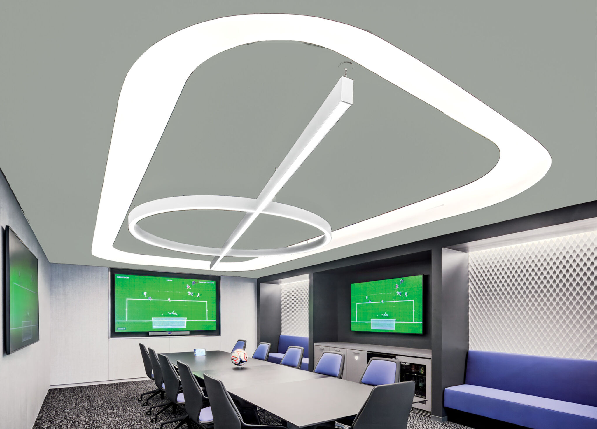 Modern conference room with large ceiling light, long table, blue chairs, wall-mounted TVs displaying a soccer game, and a blue upholstered bench along one wall.