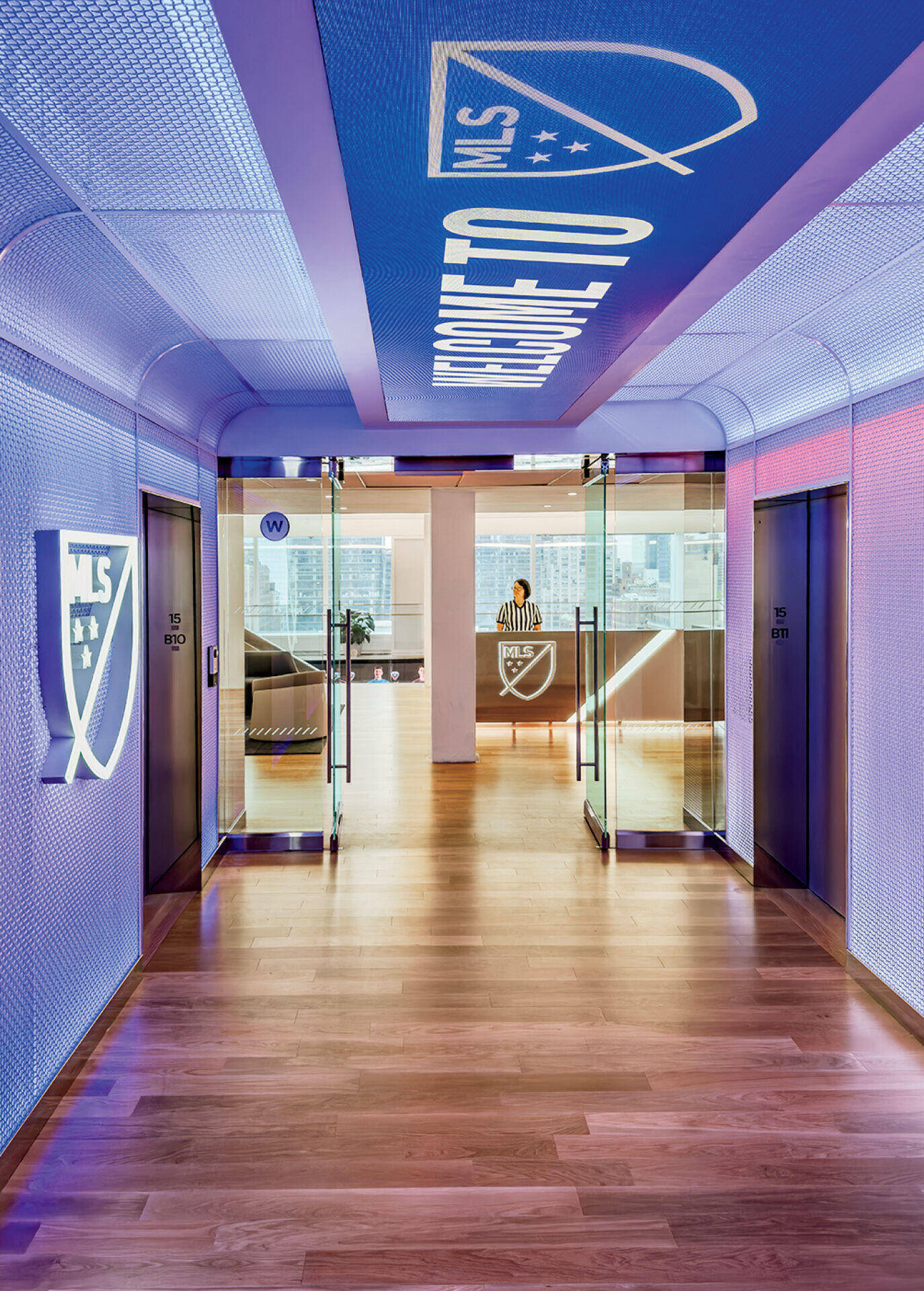 A modern hallway with wood floors, glass doors, and MLS logos on the walls leads to a reception desk and office space in the background.