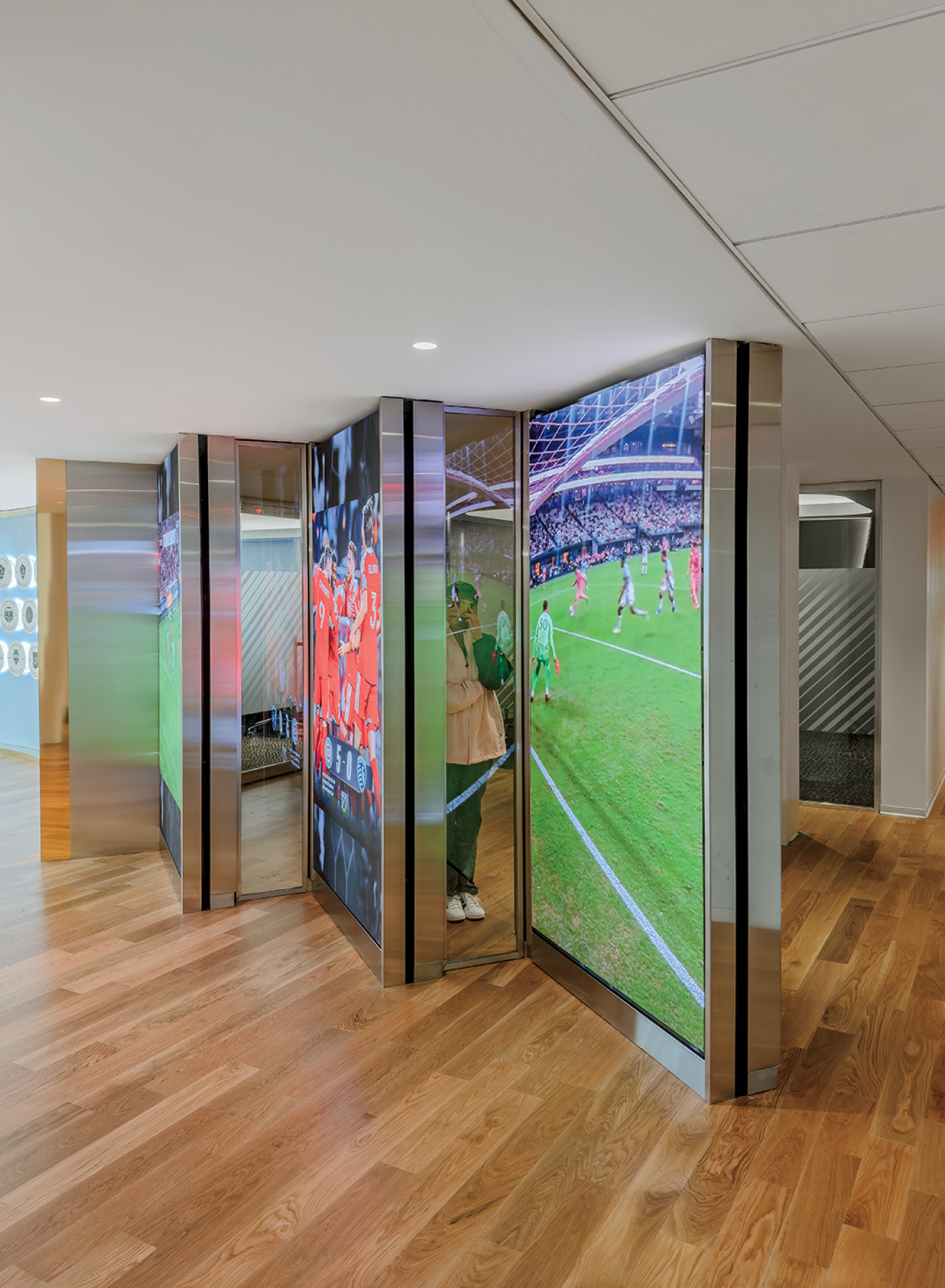 Large digital screens on angled panels display soccer images in a modern, indoor setting with wood flooring and partial glass walls.