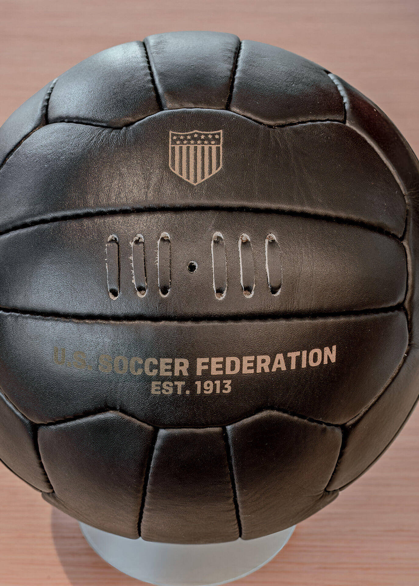 A vintage-style black leather soccer ball with the U.S. Soccer Federation crest and “U.S. Soccer Federation EST. 1913” printed on it, displayed on a stand.