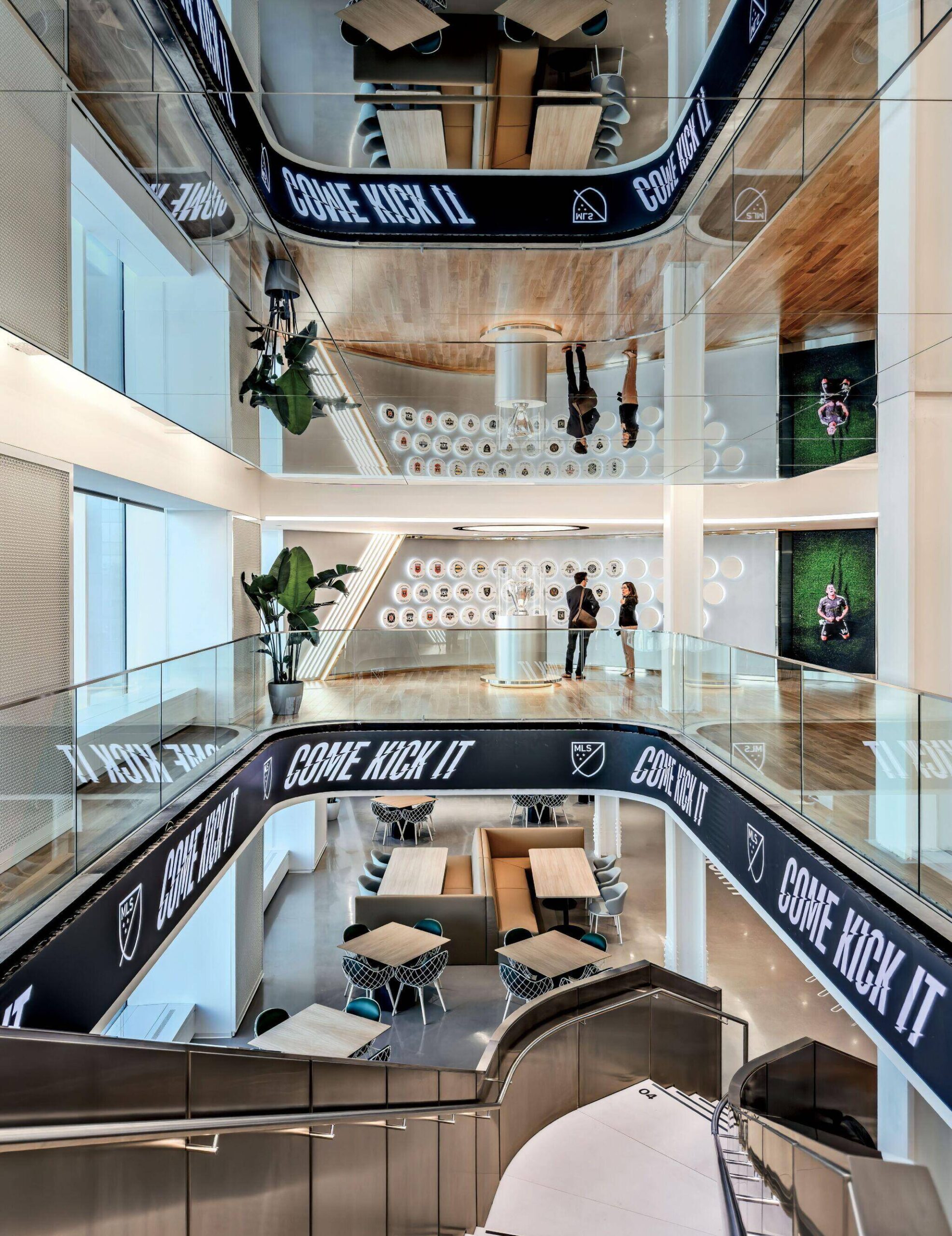 Modern multi-level interior with glass railings, mirrored ceiling, tables, chairs, large plant, and digital “COME KICK IT” banners. Two people stand on the upper level near a wall display.