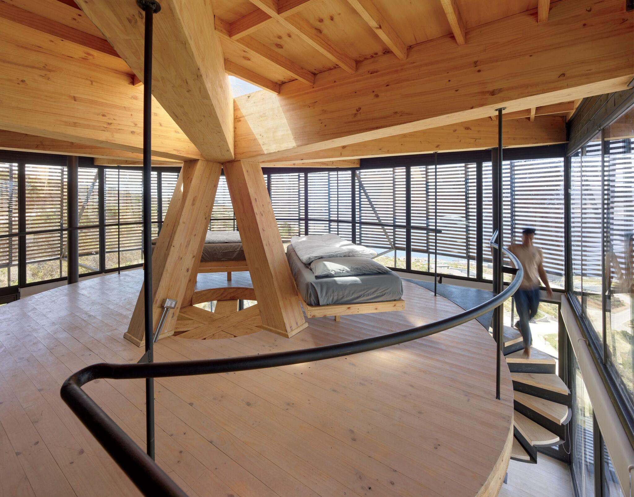 Modern wooden bedroom with large windows, spiral staircase, central wooden support structure, and platform beds; person walking upstairs.