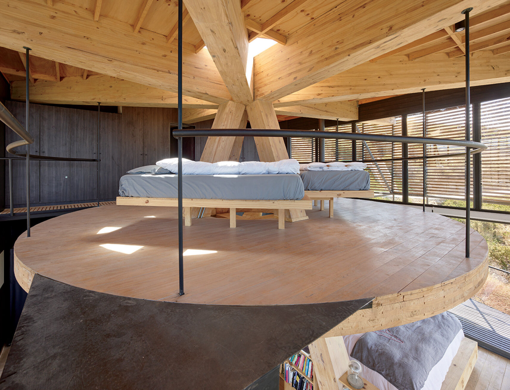 Circular loft with two beds, wood beams, and railings; natural light enters through slatted walls and ceiling opening.