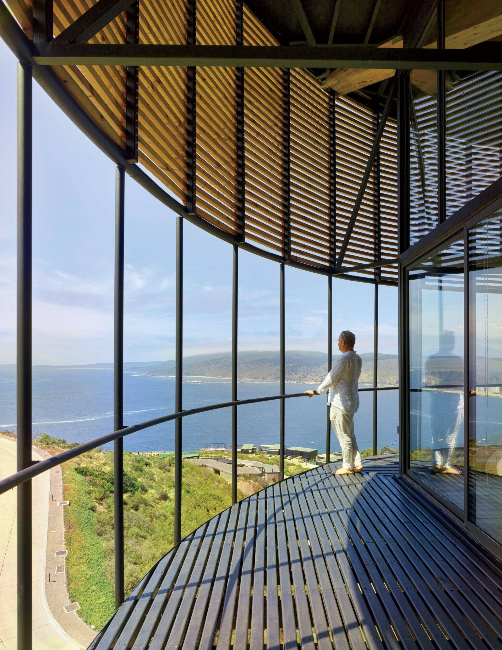 Person stands on a curved balcony with wooden slats, overlooking a coastal landscape and the sea through large floor-to-ceiling windows.