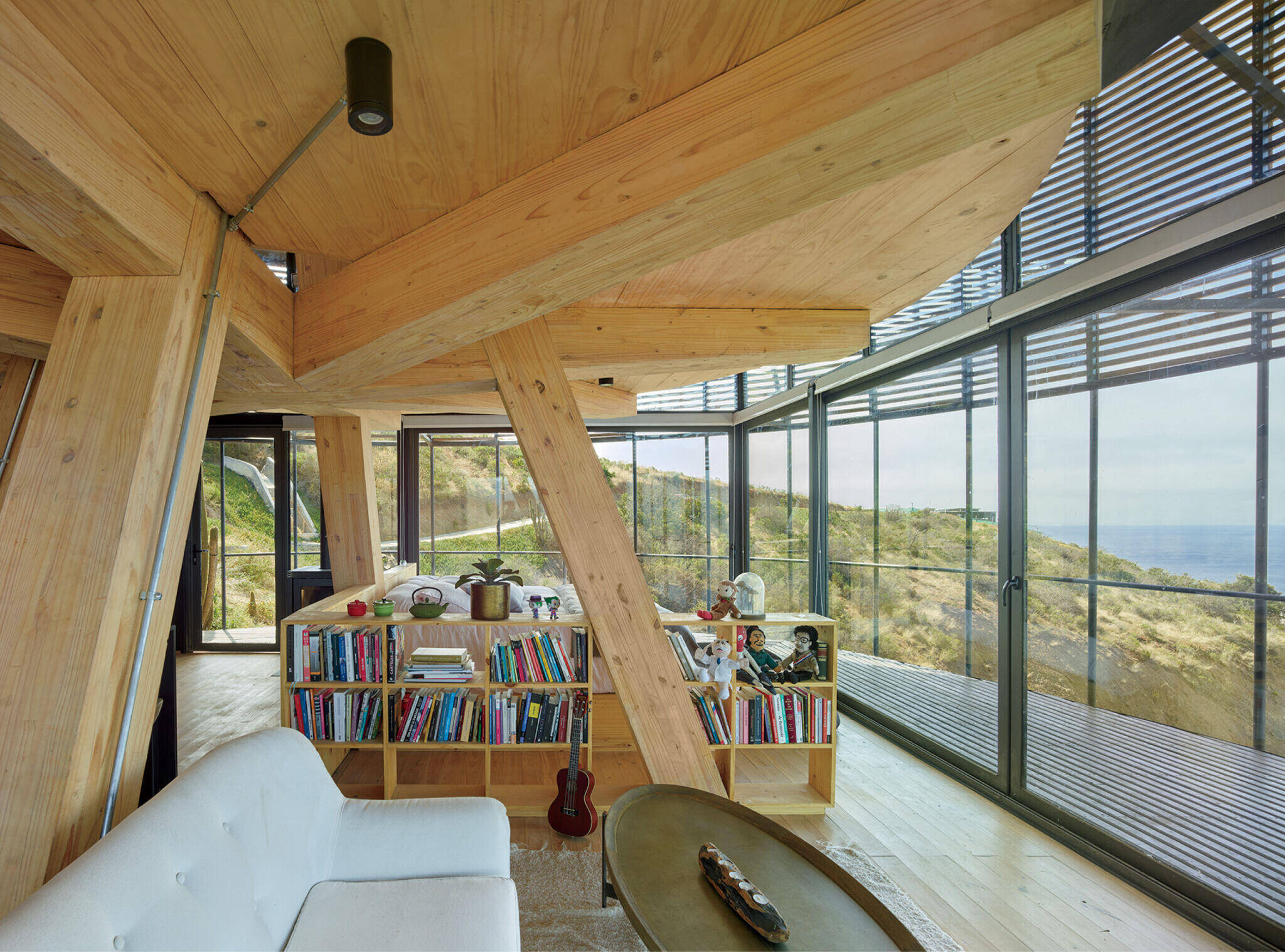 Modern wooden living room with floor-to-ceiling windows, a white couch, a low bookcase filled with books, and views of hills and ocean outside.
