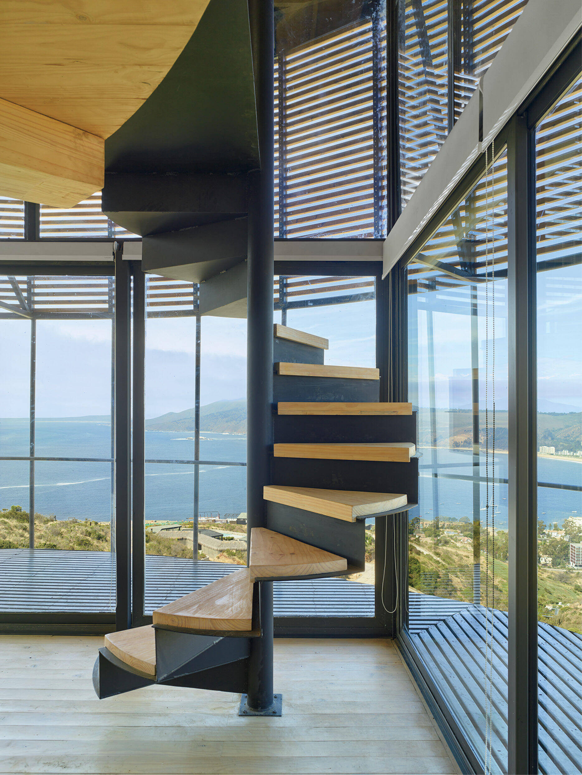 Spiral staircase with wooden steps inside a modern glass-walled room overlooking a coastline and the sea.