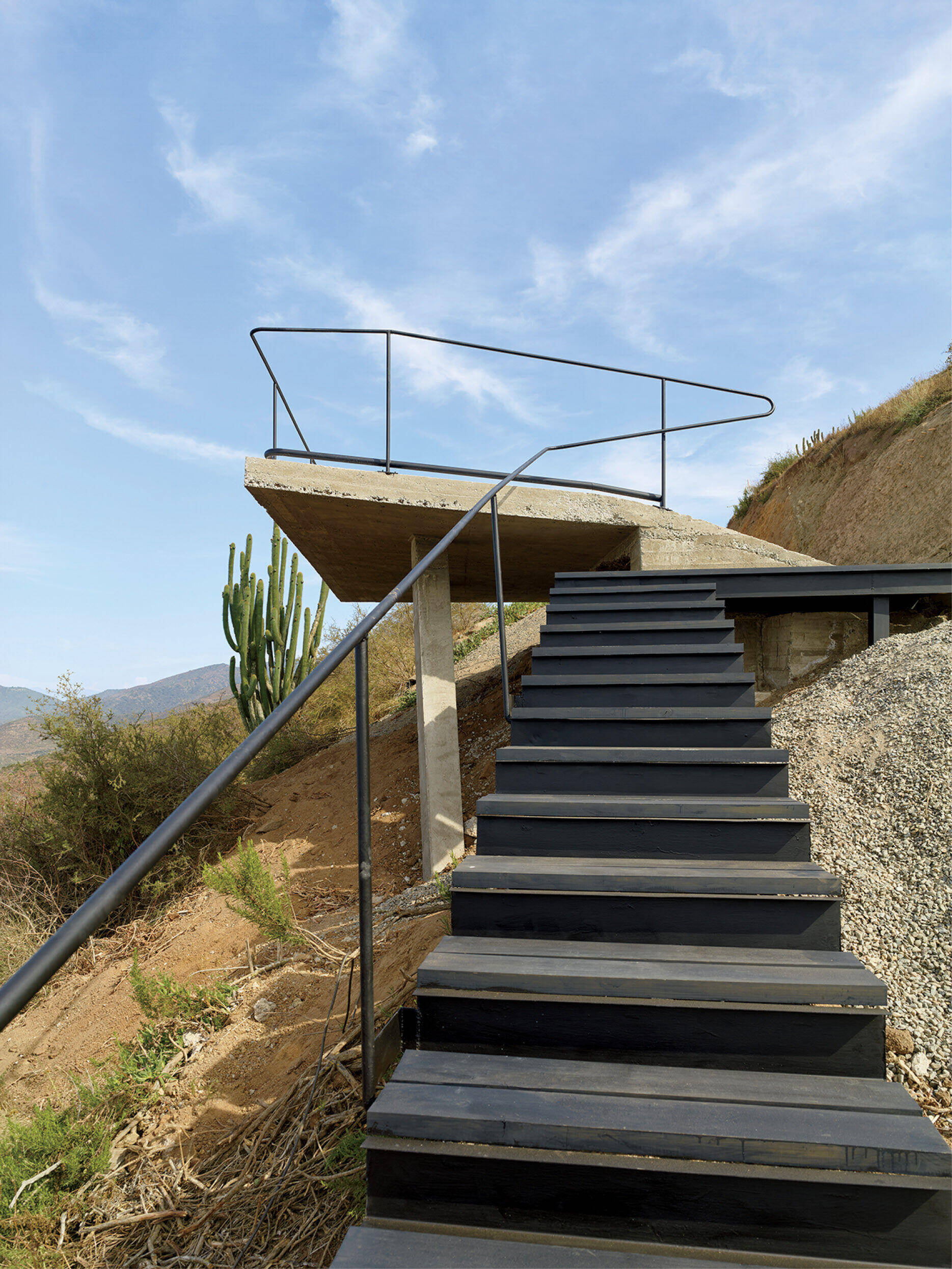 Outdoor metal staircase with a simple handrail leads up a slope to a concrete platform overlooking a desert landscape with hills and a large cactus. Blue sky and scattered clouds above.