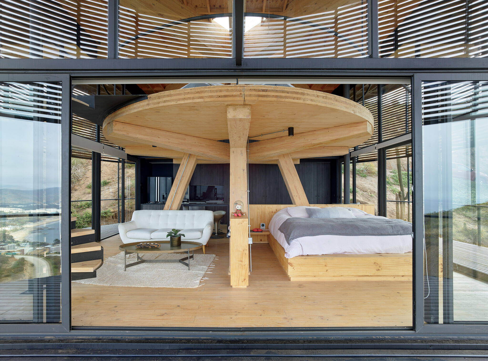 Modern bedroom with wooden beams, a bed, sofa, and coffee table, viewed through large glass doors; hills and buildings visible outside.