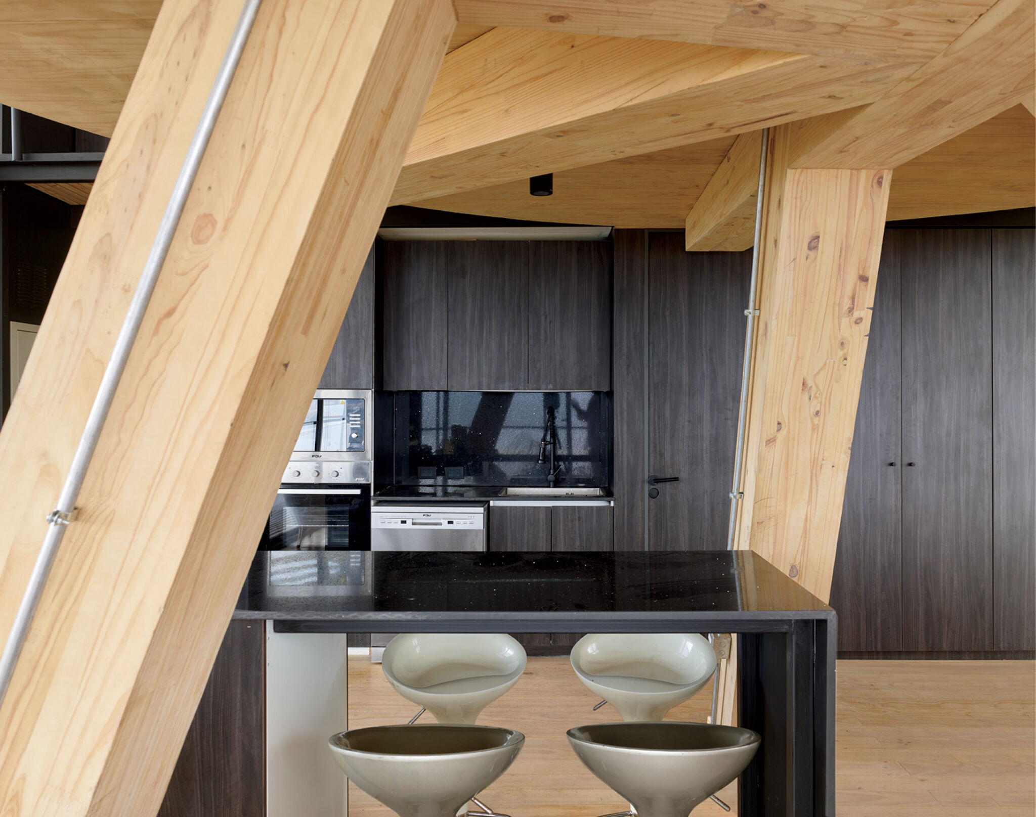 Modern kitchen with large exposed wooden beams, dark cabinets, built-in appliances, and a black countertop island with two gray bar stools.