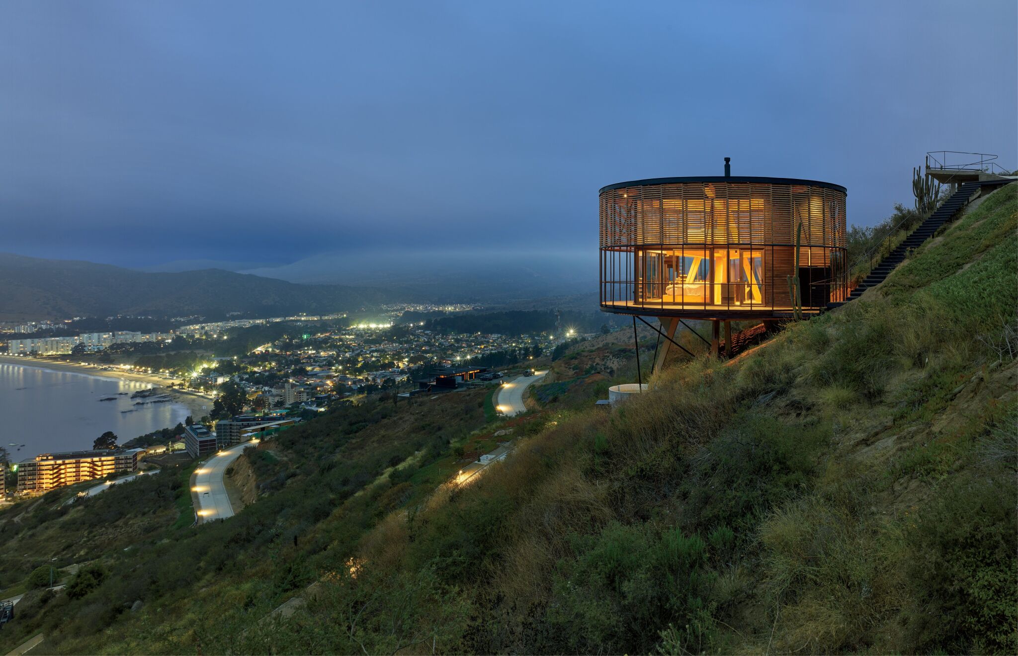 Modern circular house on stilts sits on a hillside at dusk, overlooking a coastal city with illuminated streets and buildings below.