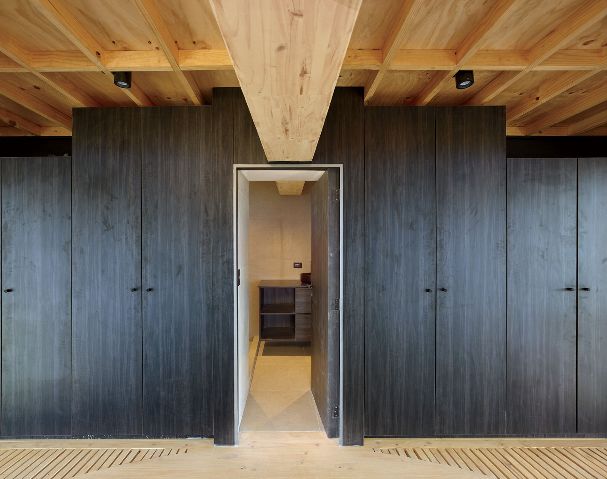 A doorway is centered in a dark wood-paneled wall, with built-in cabinets on both sides. Exposed wooden beams line the ceiling above the minimalist room.