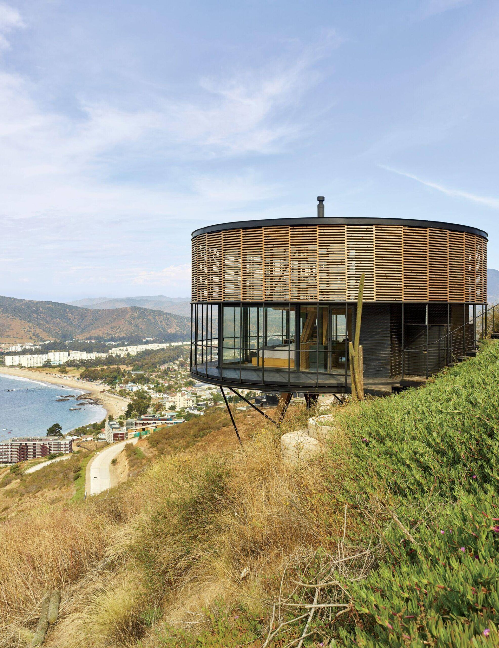Round modern house with wood slats and glass walls sits on a hillside overlooking the ocean and coastline, with distant buildings and mountains visible in the background.