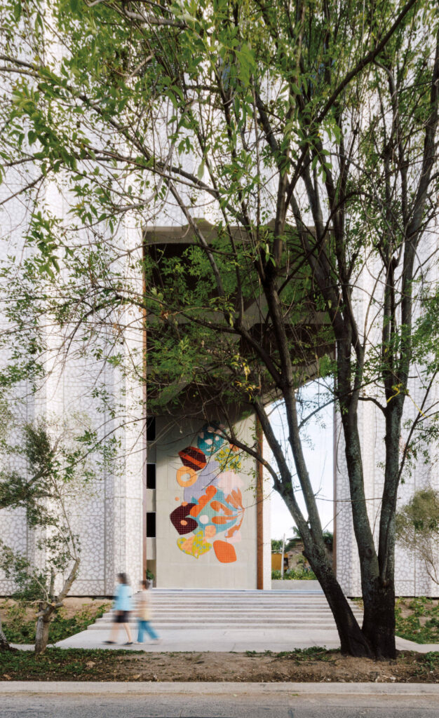 Two people walk past a modern building with a large abstract mural between tall columns, partially obscured by leafy trees.