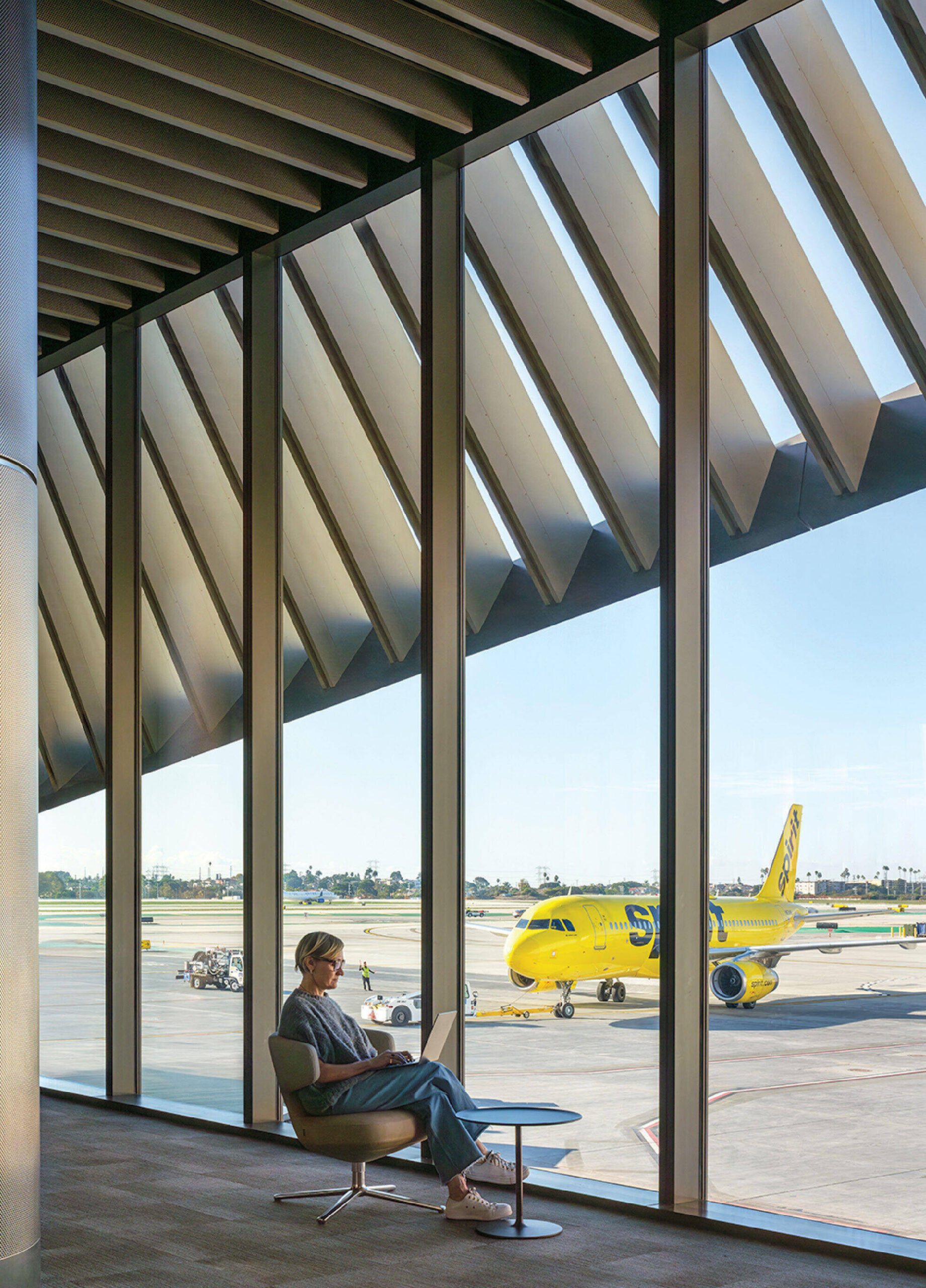 A person sits by large windows at an airport, reading, with a yellow airplane visible on the tarmac outside.