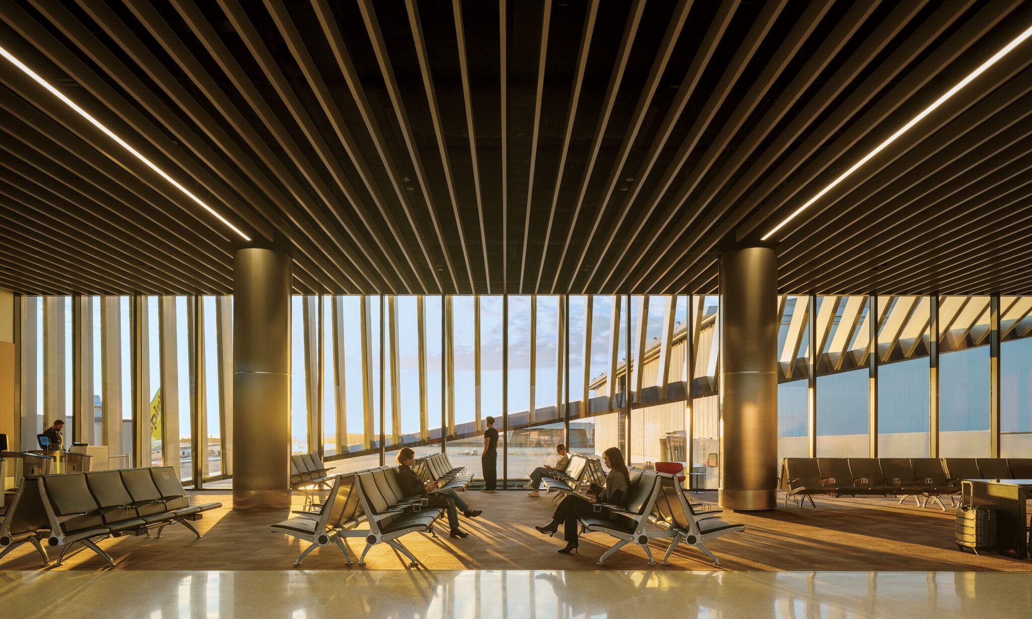 People sit and wait in an airport departure lounge with large windows, modern seating, and sunlight streaming in from outside.