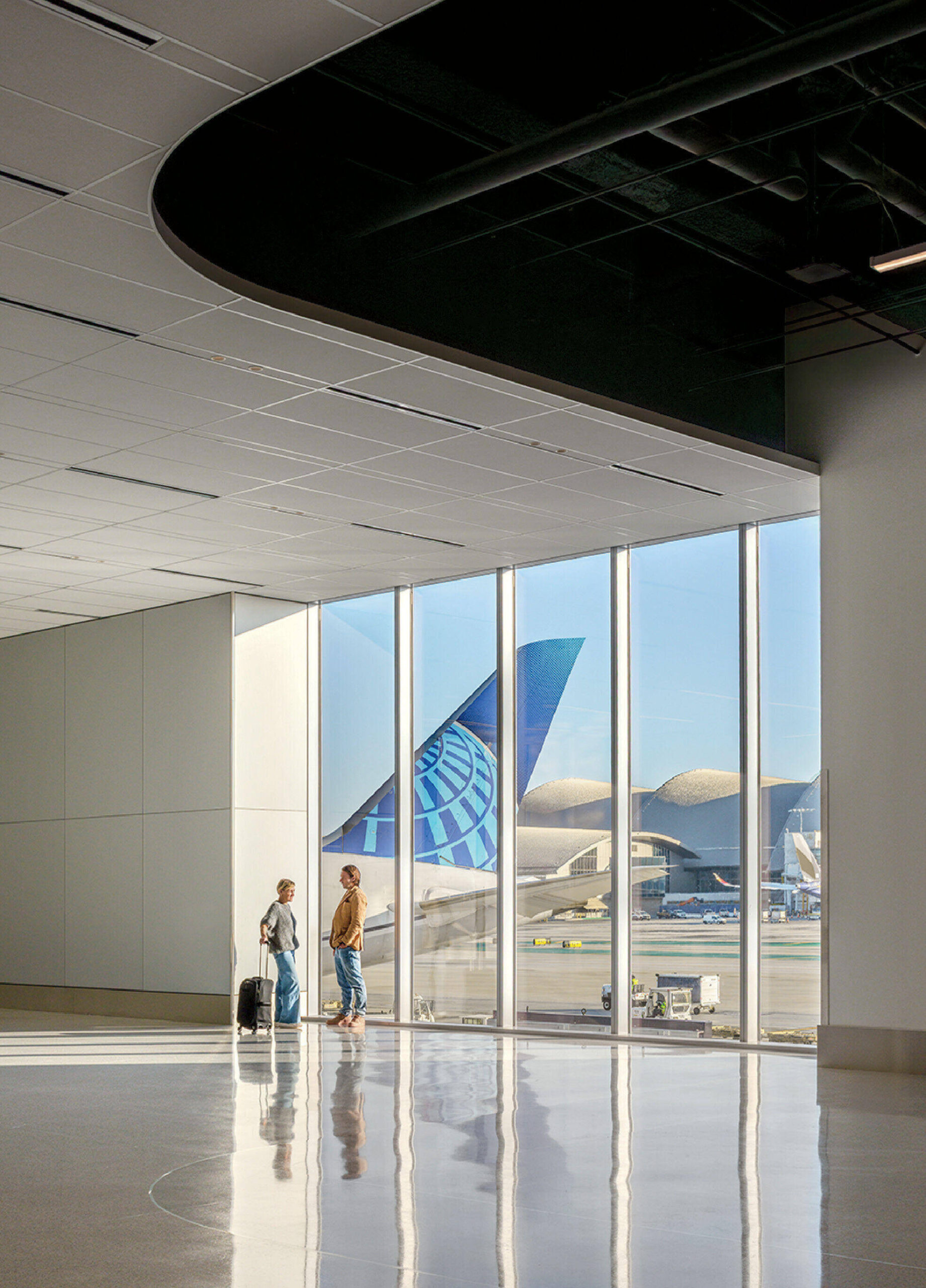 Two people with luggage stand by large airport windows, with an airplane tail and terminals visible outside on a sunny day.