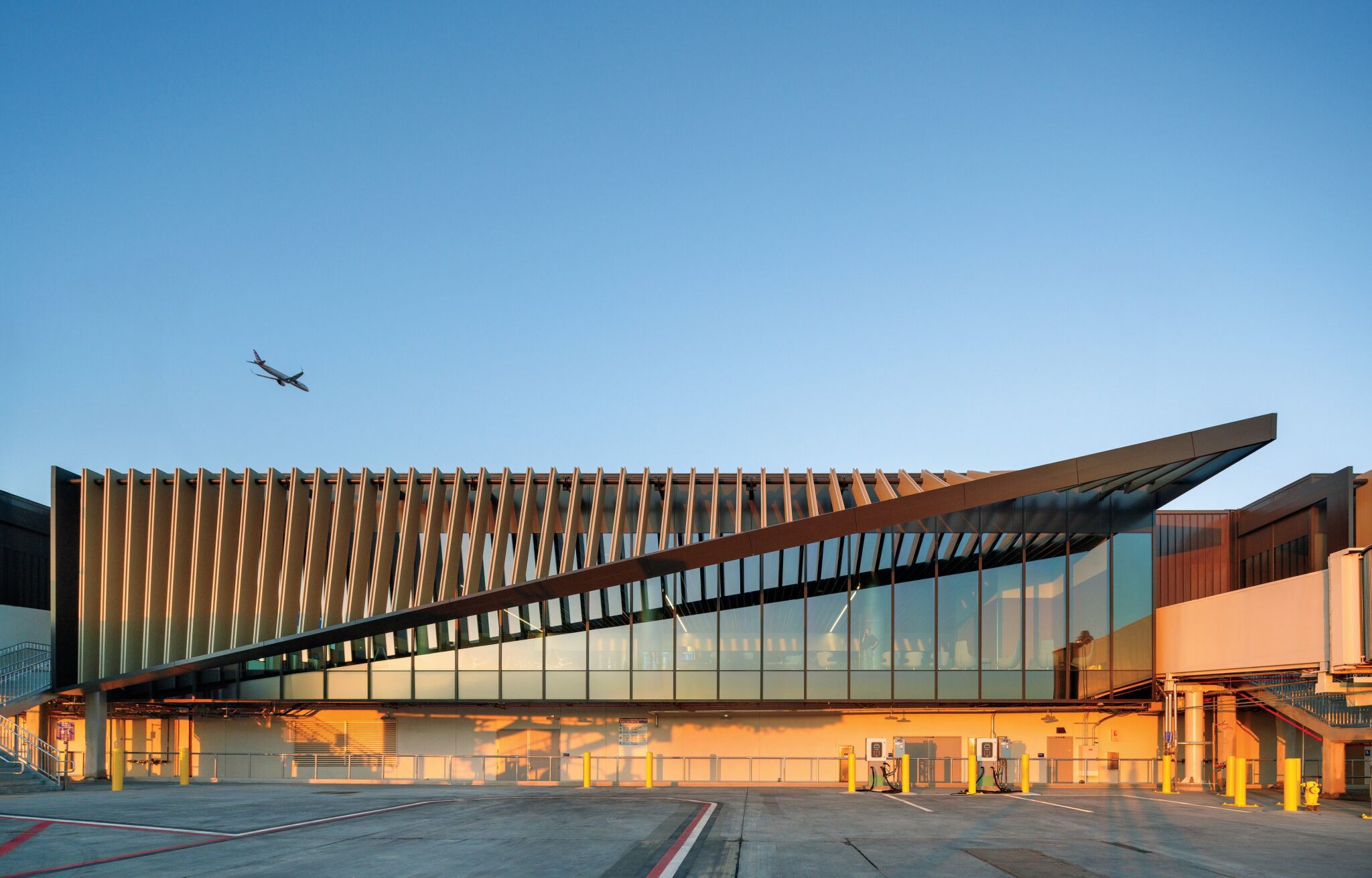 Modern airport terminal with angular roof, large glass windows, and vertical fins at sunset. An airplane is taking off in the clear blue sky above the building.