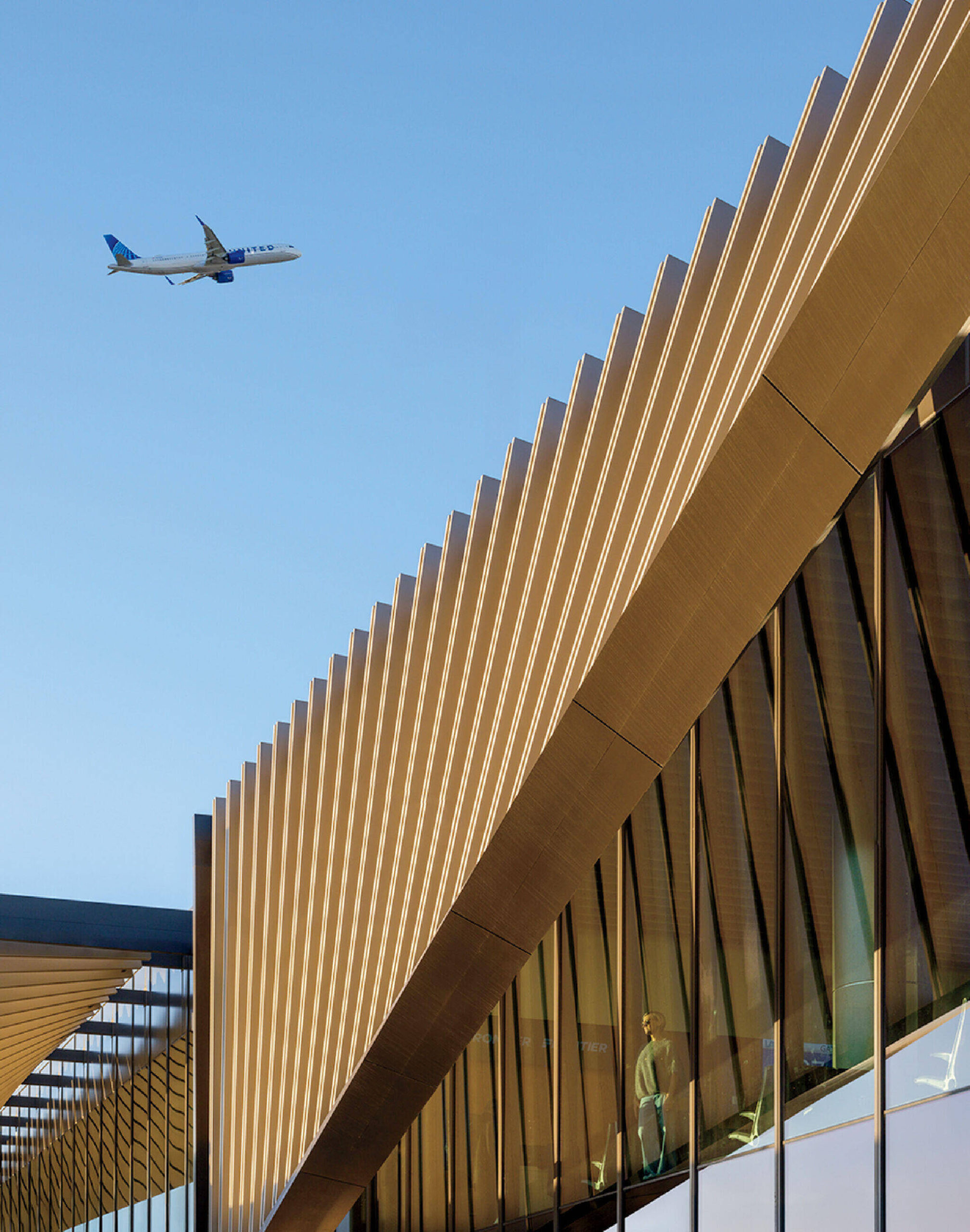 A commercial airplane flies above a modern building with angled golden panels and large glass windows under a clear blue sky.